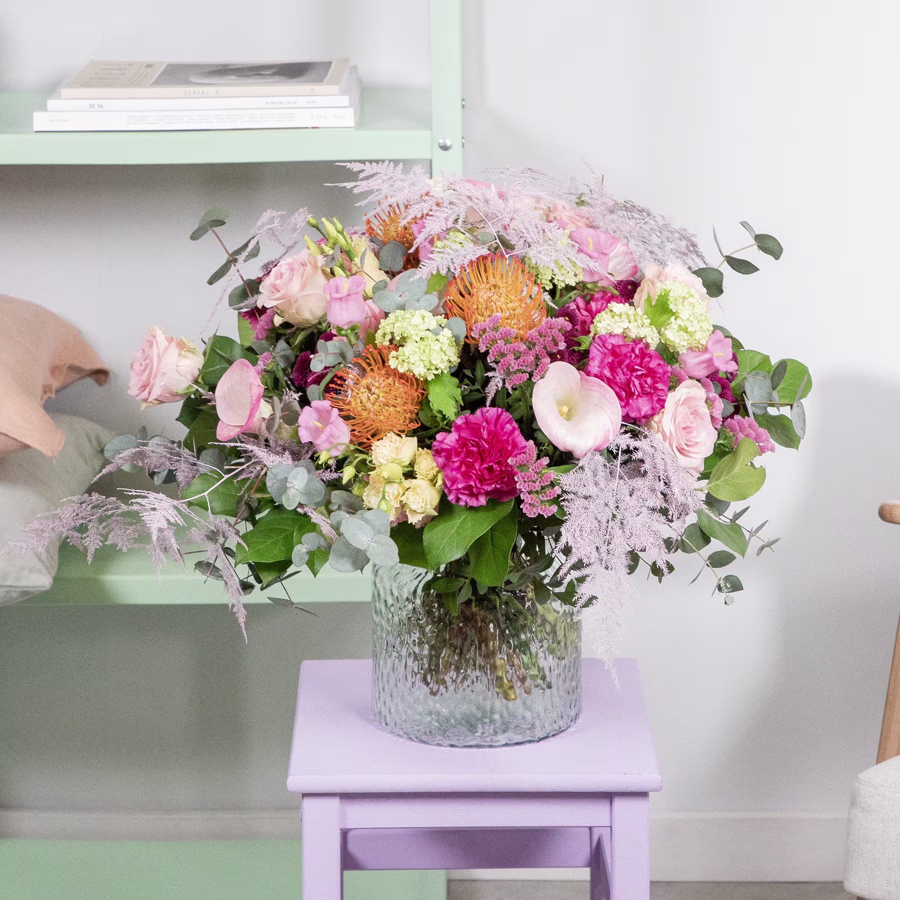 Ein leuchtender Strauß verschiedener bunter Blumen, darunter rosa Nelken, gelbe Chrysanthemen und grünes Laub, steht auf einem weißen Regal vor einer hellgrünen Wand. (Beschreibungstext KI-generiert)