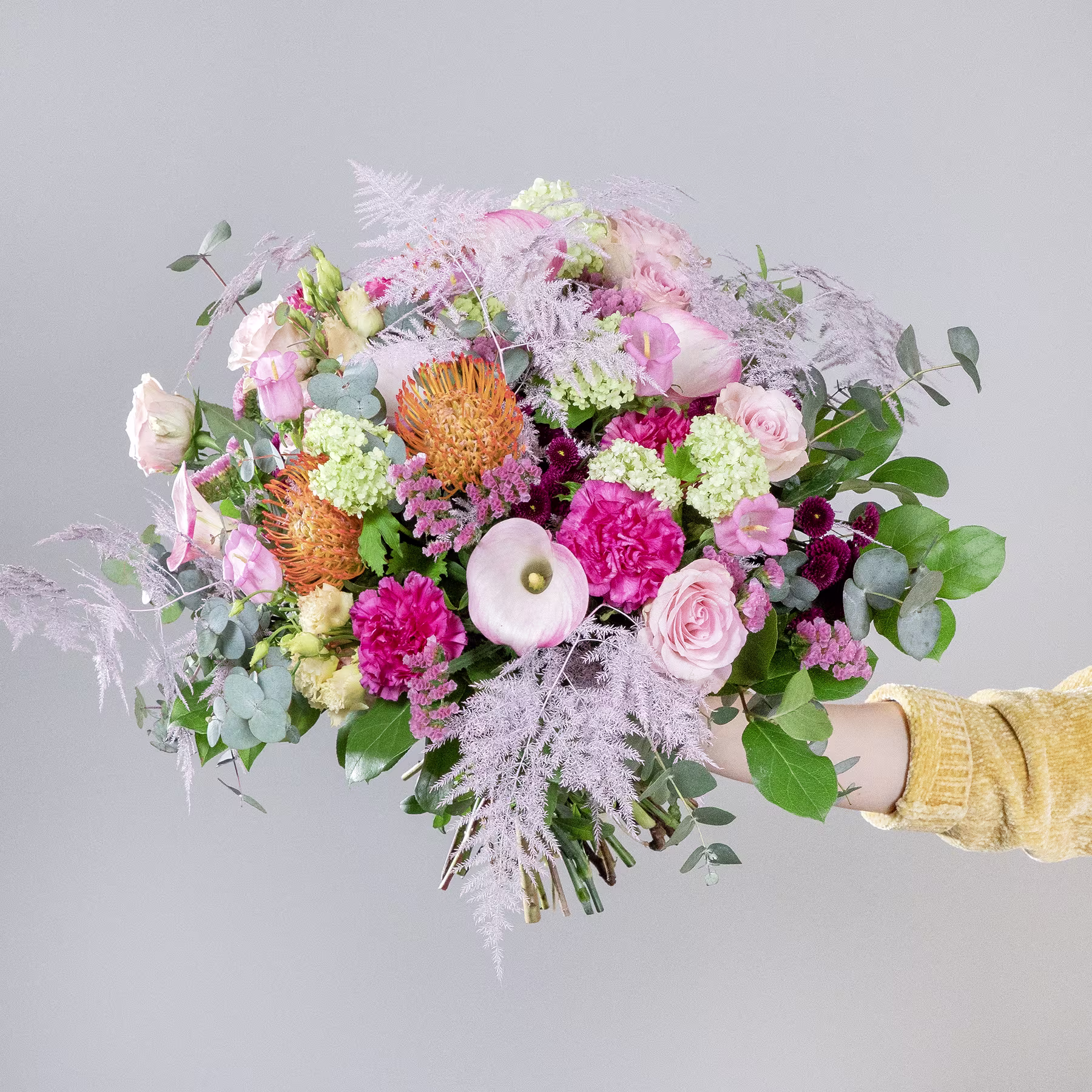 Ein lebendiger und farbenfroher Strauß verschiedener Blumen, darunter Pfingstrosen, Dahlien und Eukalyptus, den eine Person vor einem schlichten grauen Hintergrund in der Hand hält. (Beschreibungstext KI-generiert)