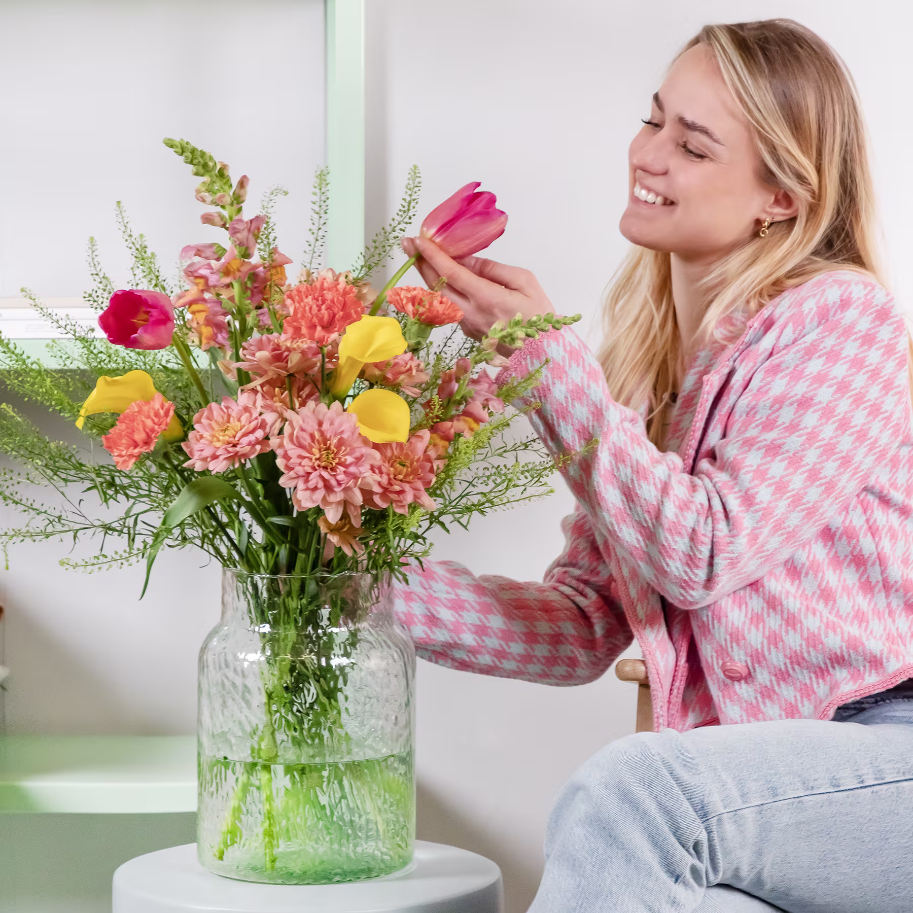 Eine lächelnde junge Frau in einem rosa-weiß karierten Pullover arrangiert vor einem hellen Hintergrund einen farbenfrohen Strauß bunter Blumen in einer Glasvase. (Beschreibungstext KI-generiert)