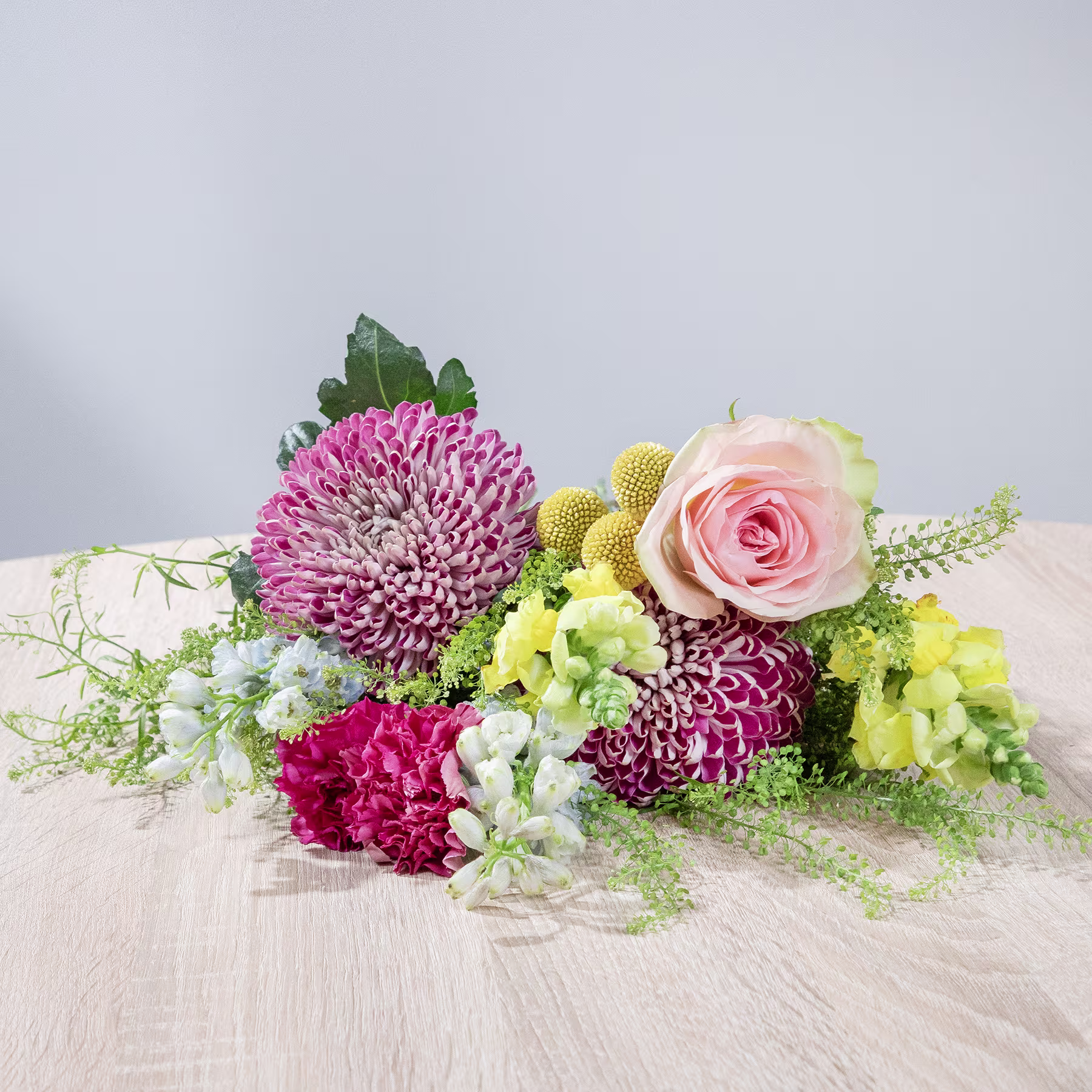 Ein farbenfroher Strauß rosa, gelber und weißer Blumen, darunter Rosen, Chrysanthemen und Grünpflanzen, liegt auf einer Holzoberfläche vor einem schlichten Hintergrund. (Beschreibungstext KI-generiert)