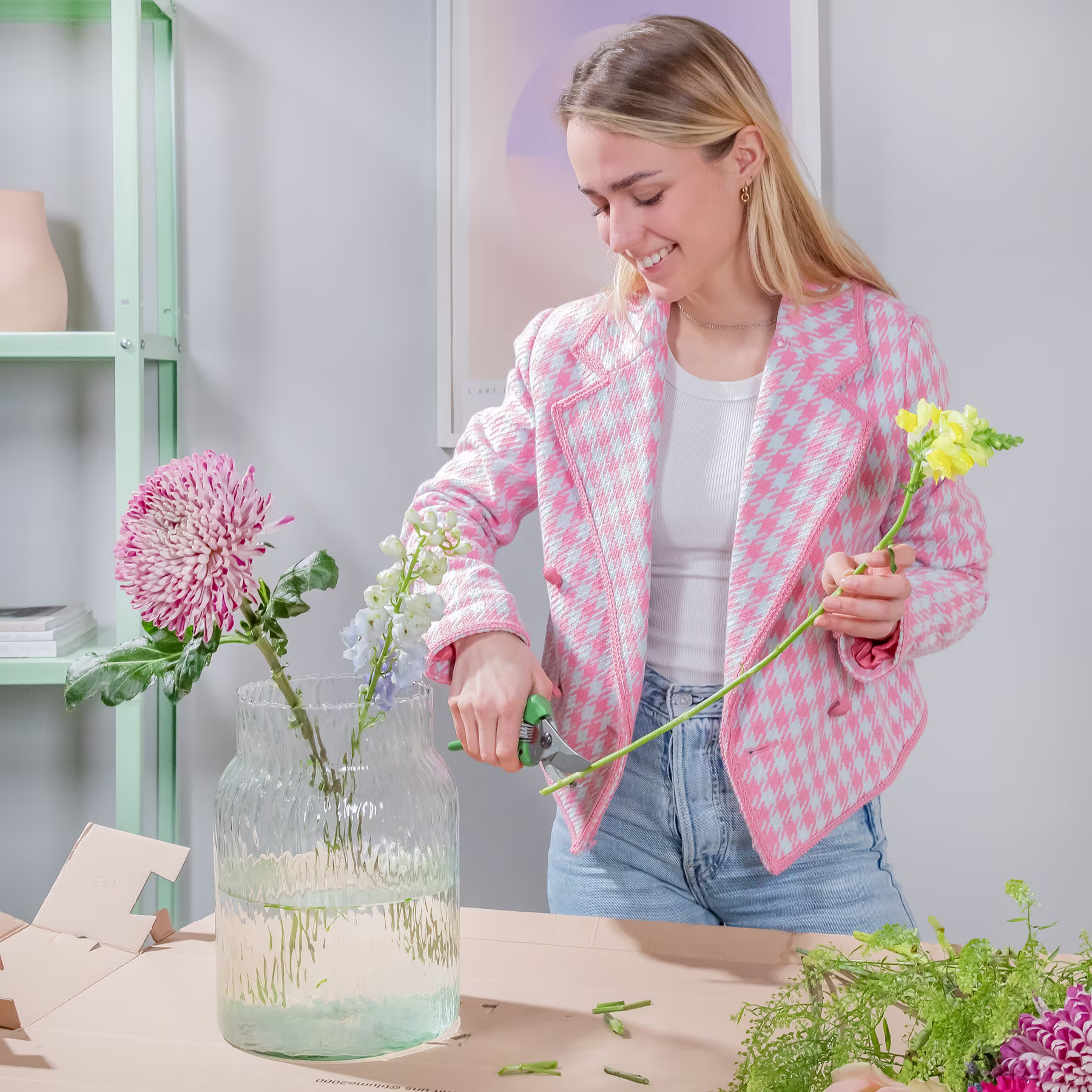 Eine junge Frau in einer rosa-weiß karierten Jacke arrangiert Blumen in einer Vase auf einem Tisch, im Hintergrund ist ein grünes Regal zu sehen. (Beschreibungstext KI-generiert)