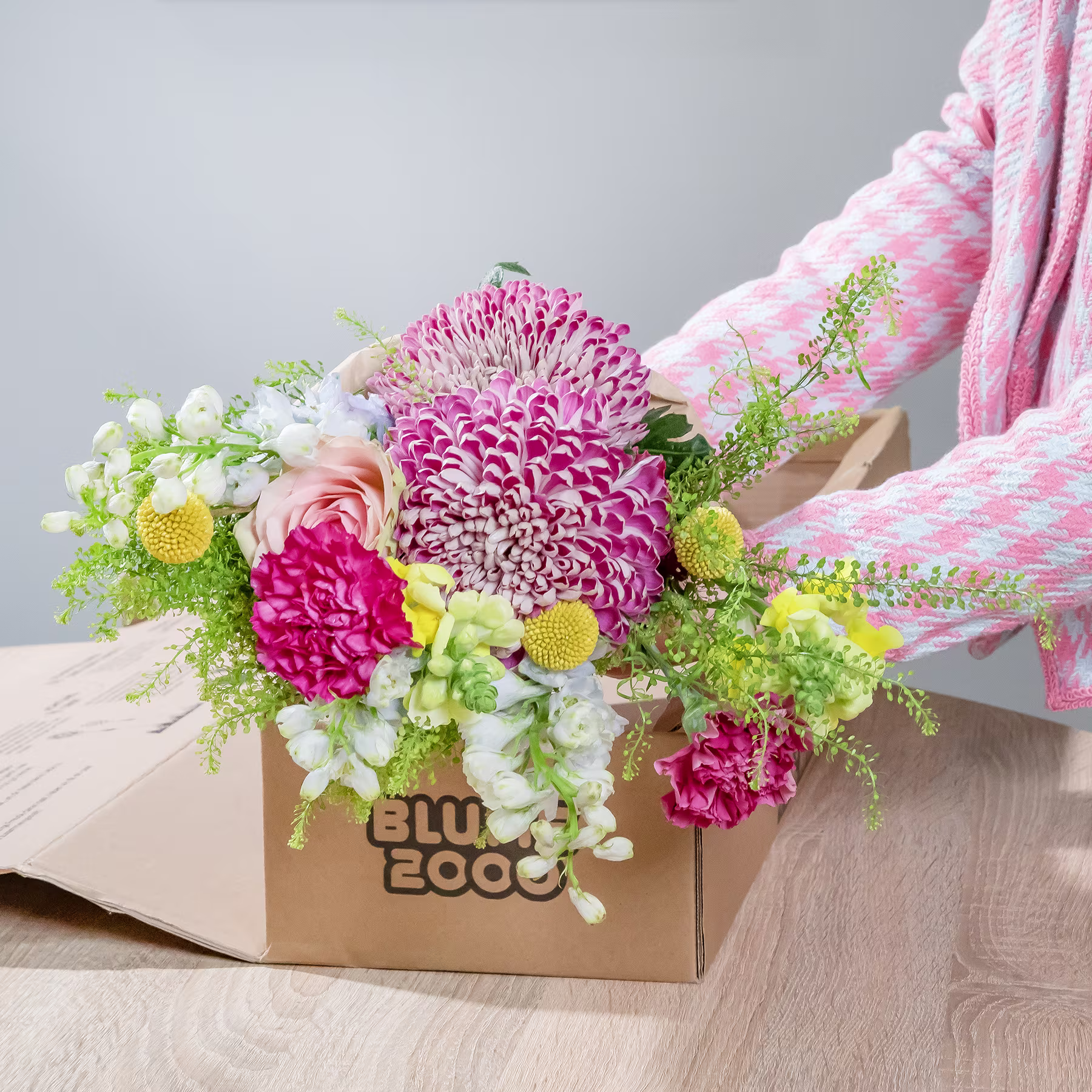 Ein lebendiger und farbenfroher Strauß verschiedener Blumen, darunter rosa Chrysanthemen, weiße Nelken und gelbe Gänseblümchen, arrangiert in einer Holzkiste auf einer Holzoberfläche. (Beschreibungstext KI-generiert)