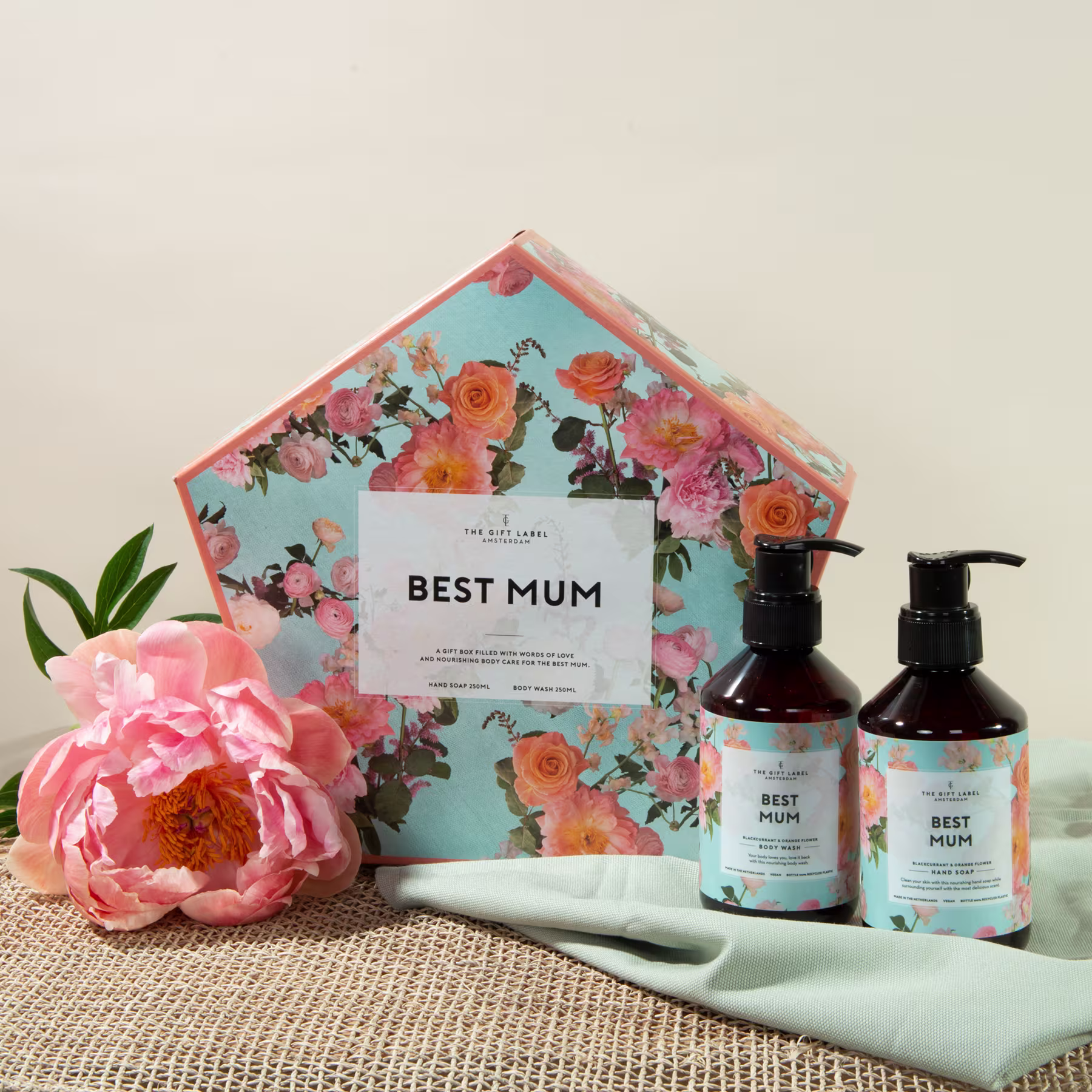 Ein Umschlag mit Blumenmuster, der eine „Beste Mama“-Karte enthält, umgeben von einer rosa Pfingstrose, einem grünen Pflanzenblatt und zwei Flaschen mit scheinbar Handseife oder Lotion. (Beschreibungstext KI-generiert)