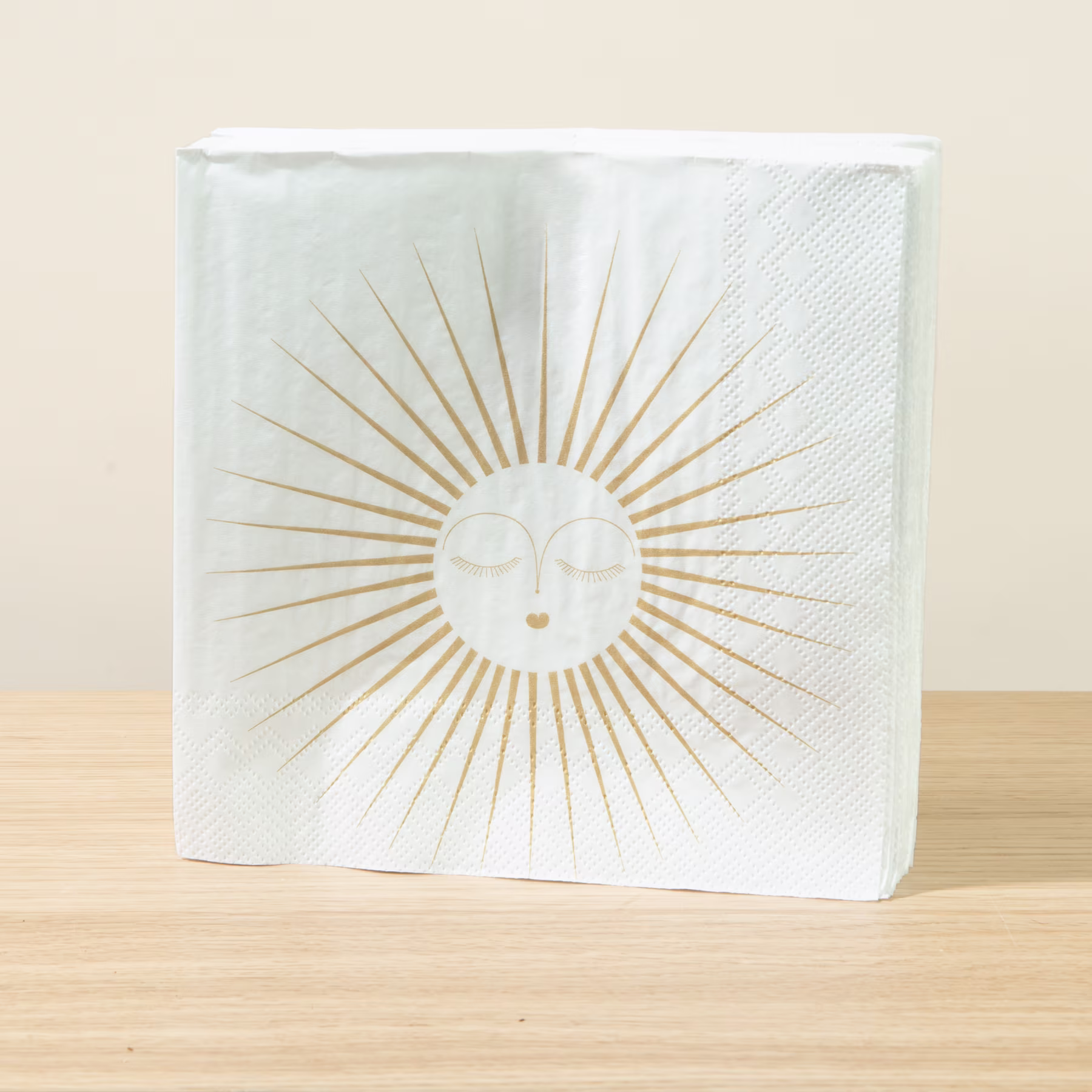 Eine weiße quadratische Serviette mit einem goldenen sonnenähnlichen Design und einem stilisierten Gesicht in der Mitte, die auf einer Holzoberfläche liegt. (Beschreibungstext KI-generiert)