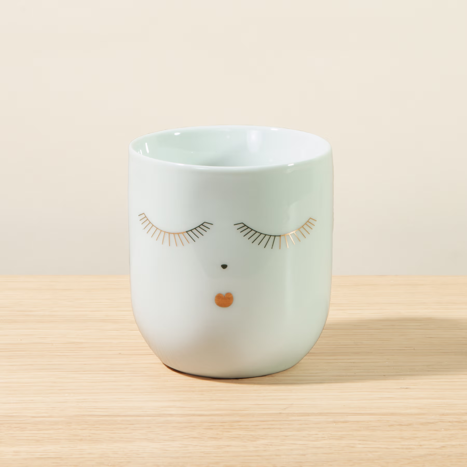 Eine weiße Keramiktasse oder ein Becher mit einem einfachen Gesichtsdesign, mit geschlossenen Augen und einem kleinen orangefarbenen Punkt als Nase, steht auf einer Holzoberfläche vor einem hellen Hintergrund. (Beschreibungstext KI-generiert)