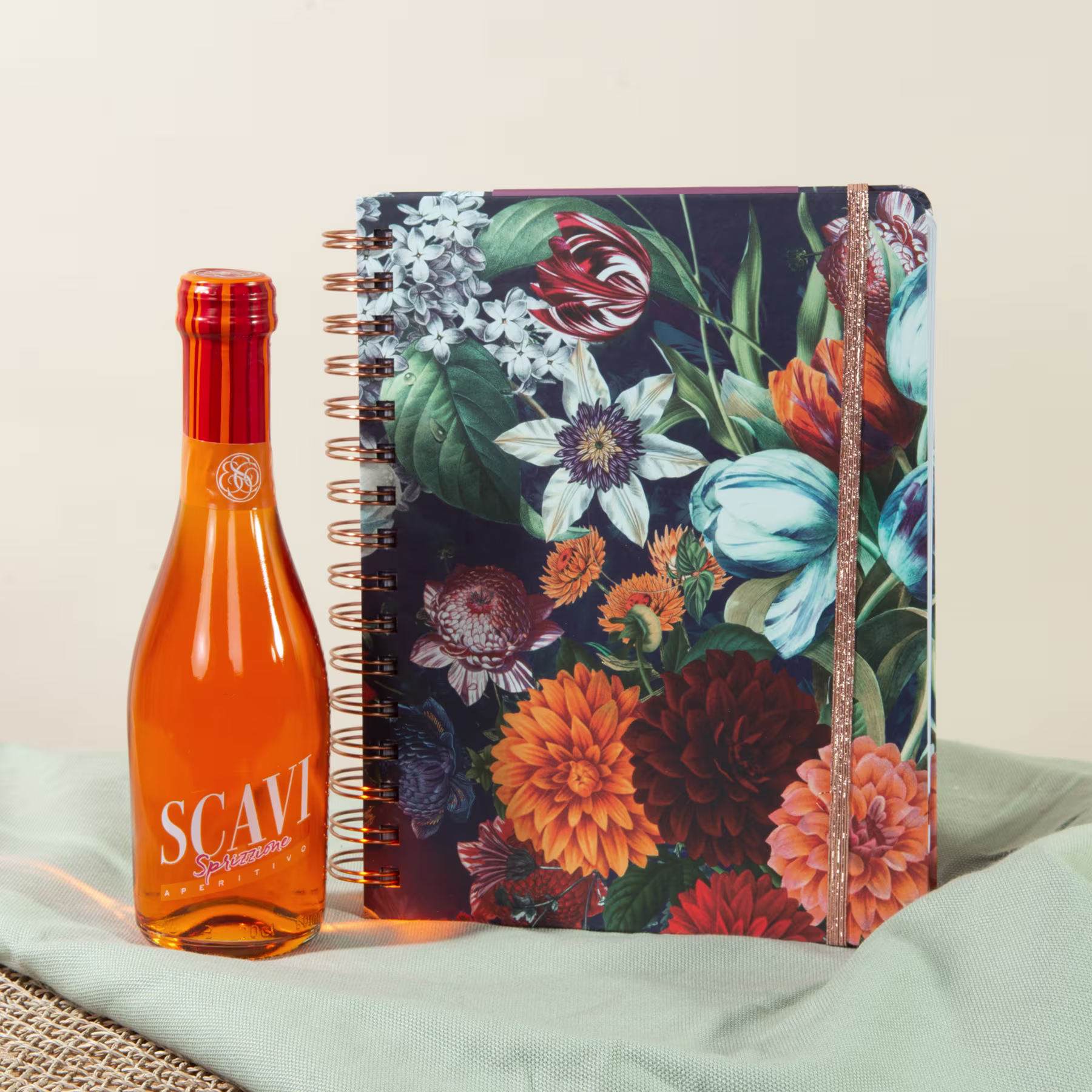 Auf einem hellen Stoffhintergrund sind ein farbenfrohes Notizbuch mit Blumenmuster und eine Flasche mit orangefarbener Flüssigkeit abgebildet. (Beschreibungstext KI-generiert)