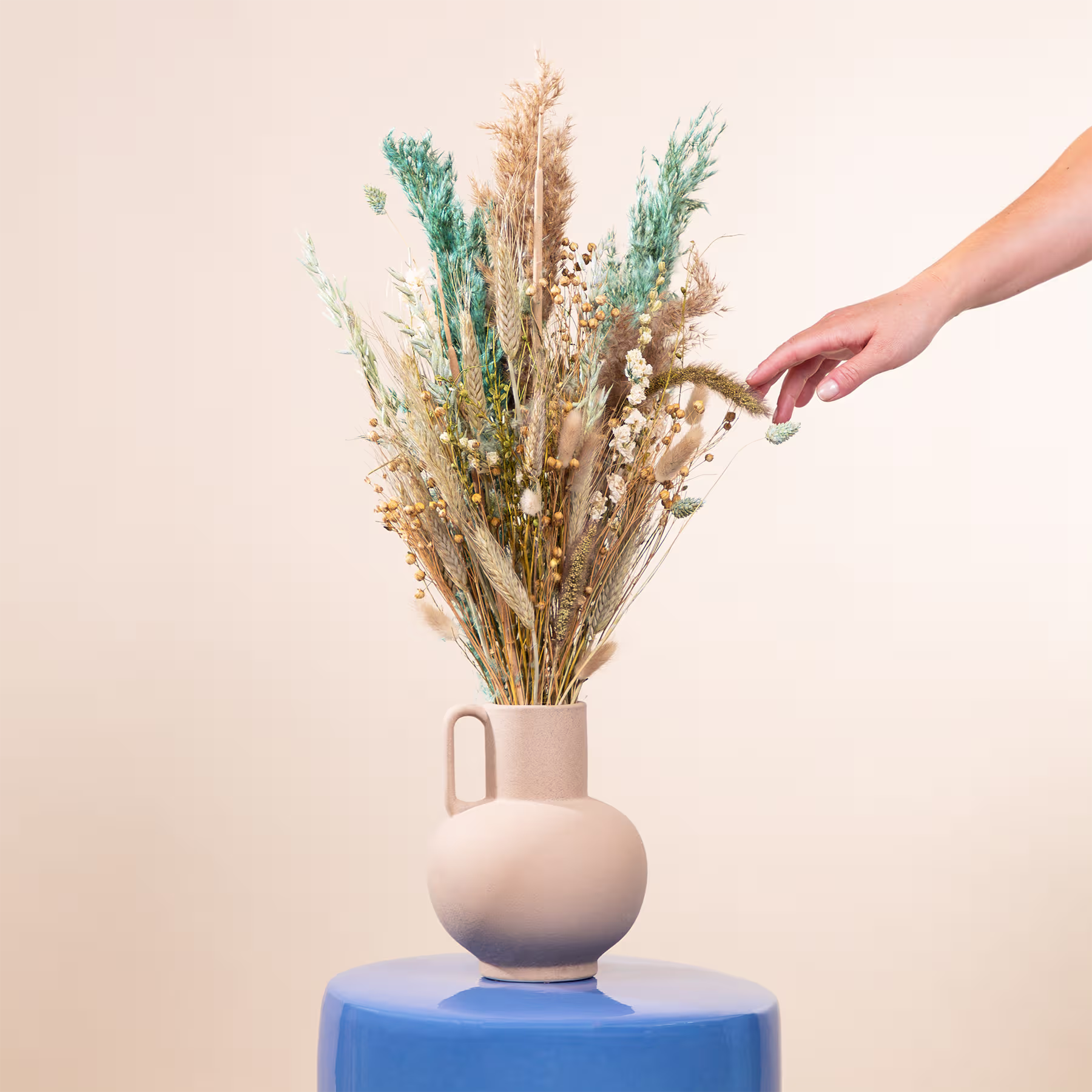 Eine Hand arrangiert einen Strauß getrockneter Blumen und Pflanzen in einer beigen Keramikvase, die auf einer blauen Fläche vor einem neutralen Hintergrund steht. (Beschreibungstext KI-generiert)