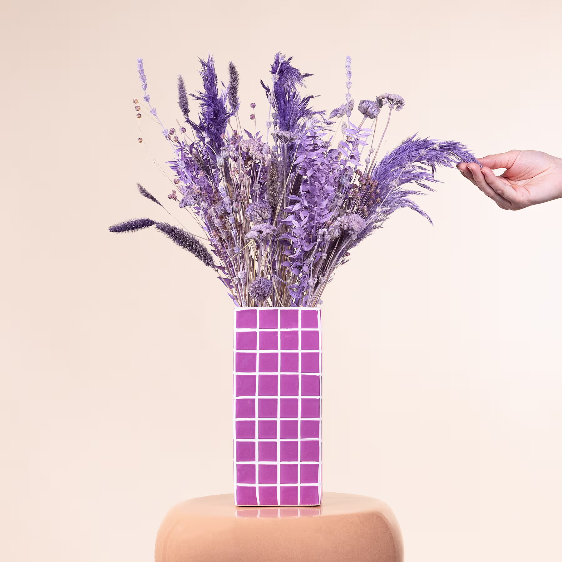 Eine Hand hält einen leuchtenden Strauß lila Blumen in einer Vase mit einem rosa-weißen Schachbrettmuster vor einem neutralen Hintergrund. (Beschreibungstext KI-generiert)