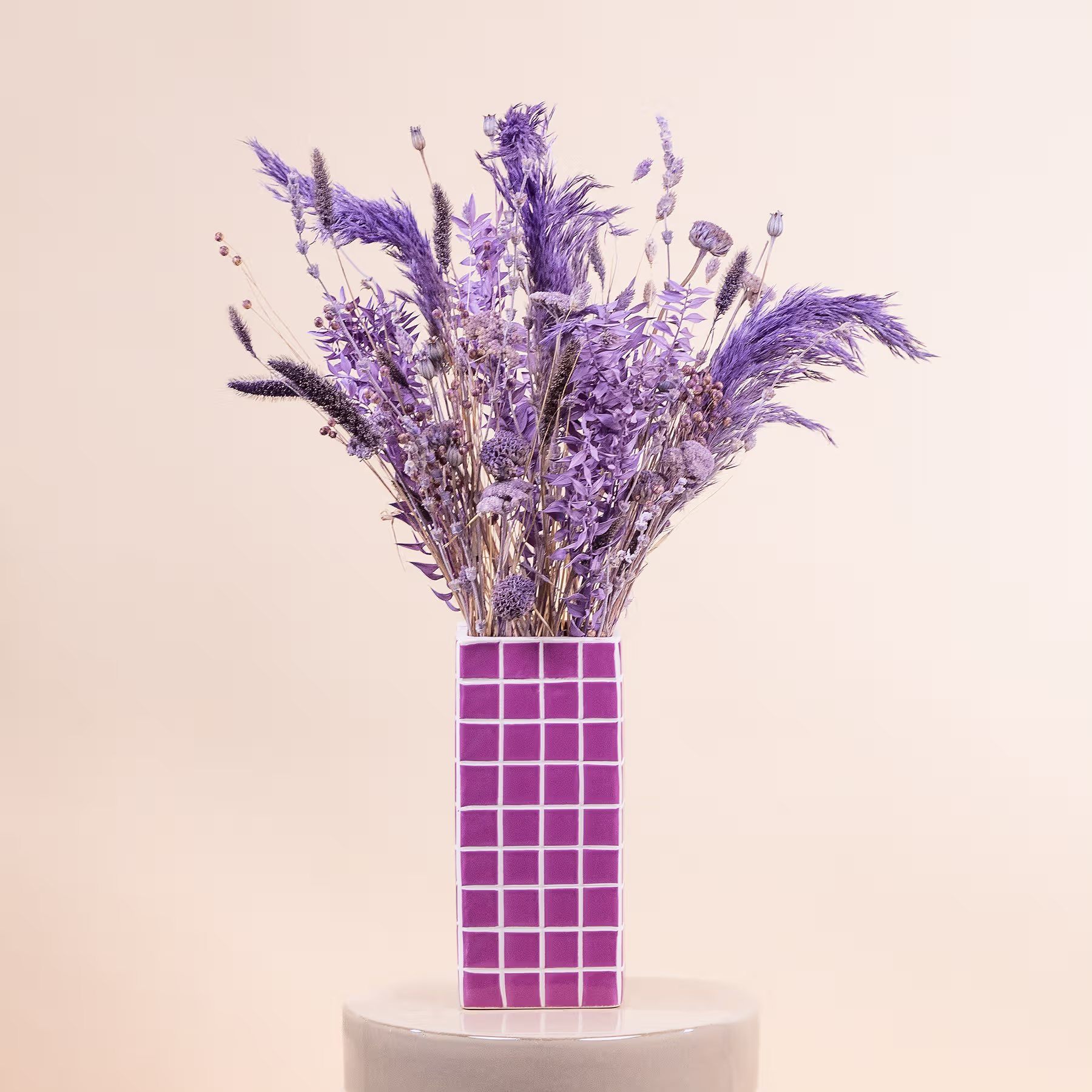 Ein farbenfroher Strauß violetter Blumen, darunter Lavendel und andere zarte Blüten, wird in einer geometrischen rosa Vase vor einem neutralen beigen Hintergrund präsentiert. (Beschreibungstext KI-generiert)