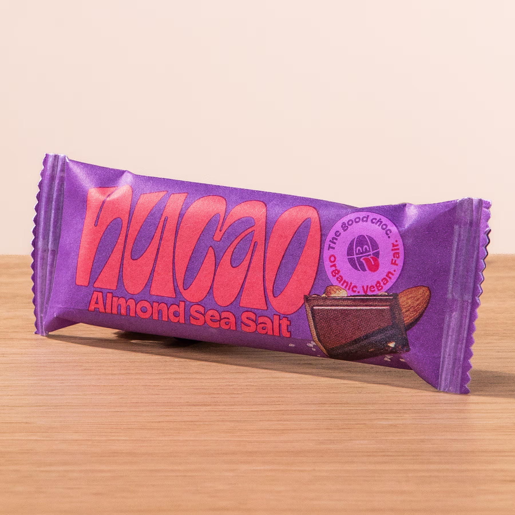 Eine lilafarbene Schokoriegelverpackung mit dem Wort „Nakao“ und der Geschmacksrichtung „Mandel-Meersalz“, die deutlich sichtbar auf einer Holzoberfläche abgebildet ist. (Beschreibungstext KI-generiert)