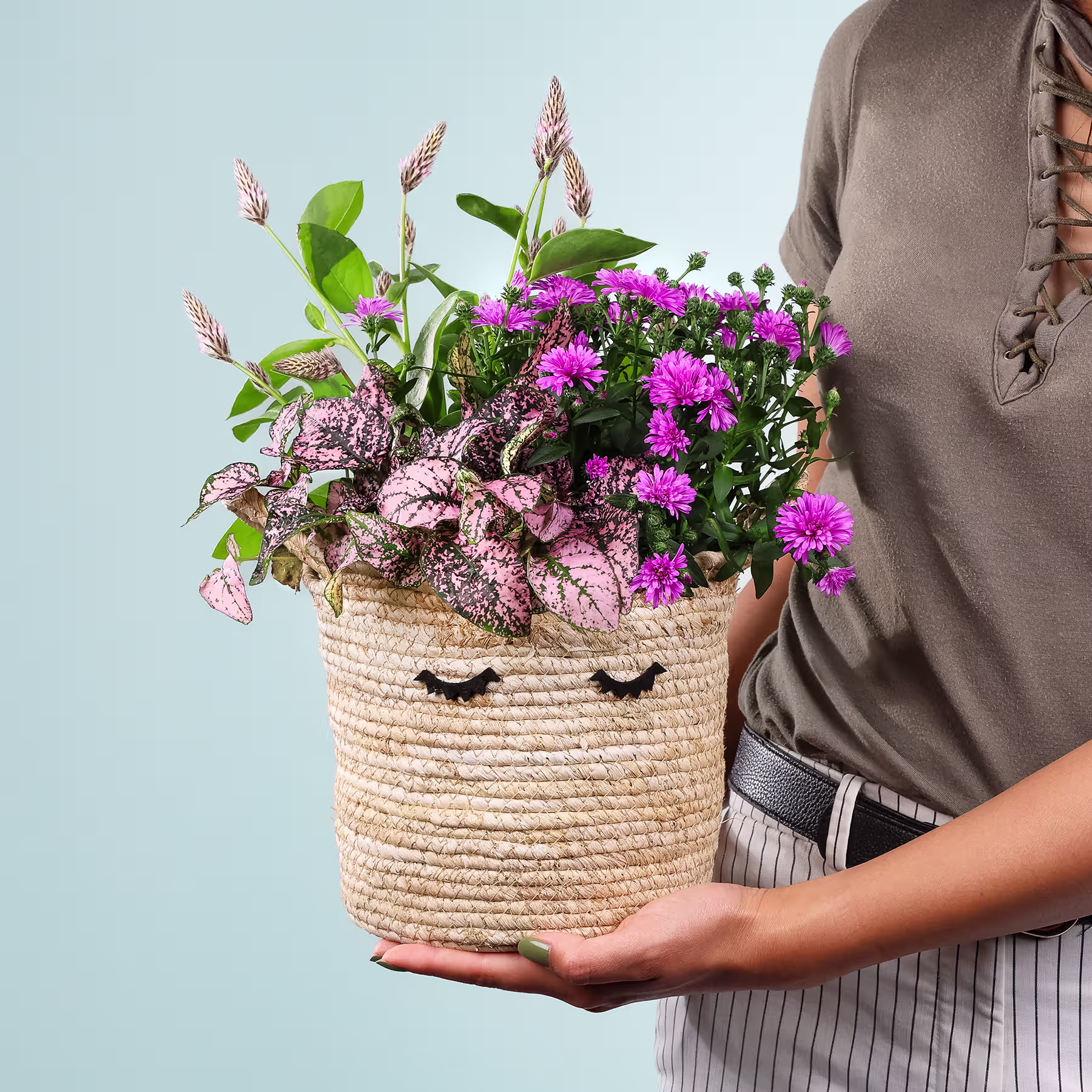 Eine Person hält einen geflochtenen Korb voller leuchtend rosa und violetter Blumen vor einem hellblauen Hintergrund. (Beschreibungstext KI-generiert)