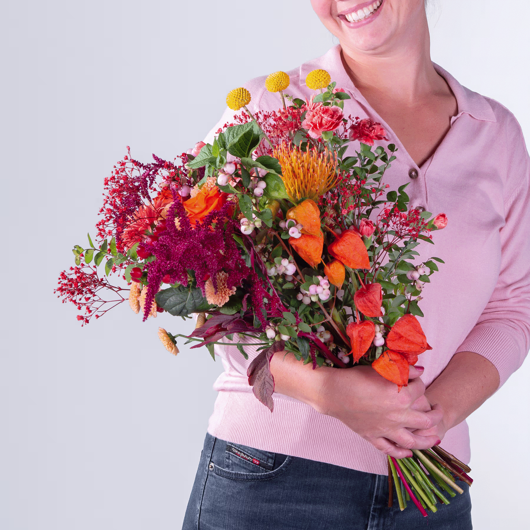 Ein leuchtender Strauß bunter Blumen, darunter Proteas, Chrysanthemen und andere Blüten, wird von einer lächelnden Person in einem rosa Oberteil vor einem schlichten Hintergrund gehalten. (Beschreibungstext KI-generiert)