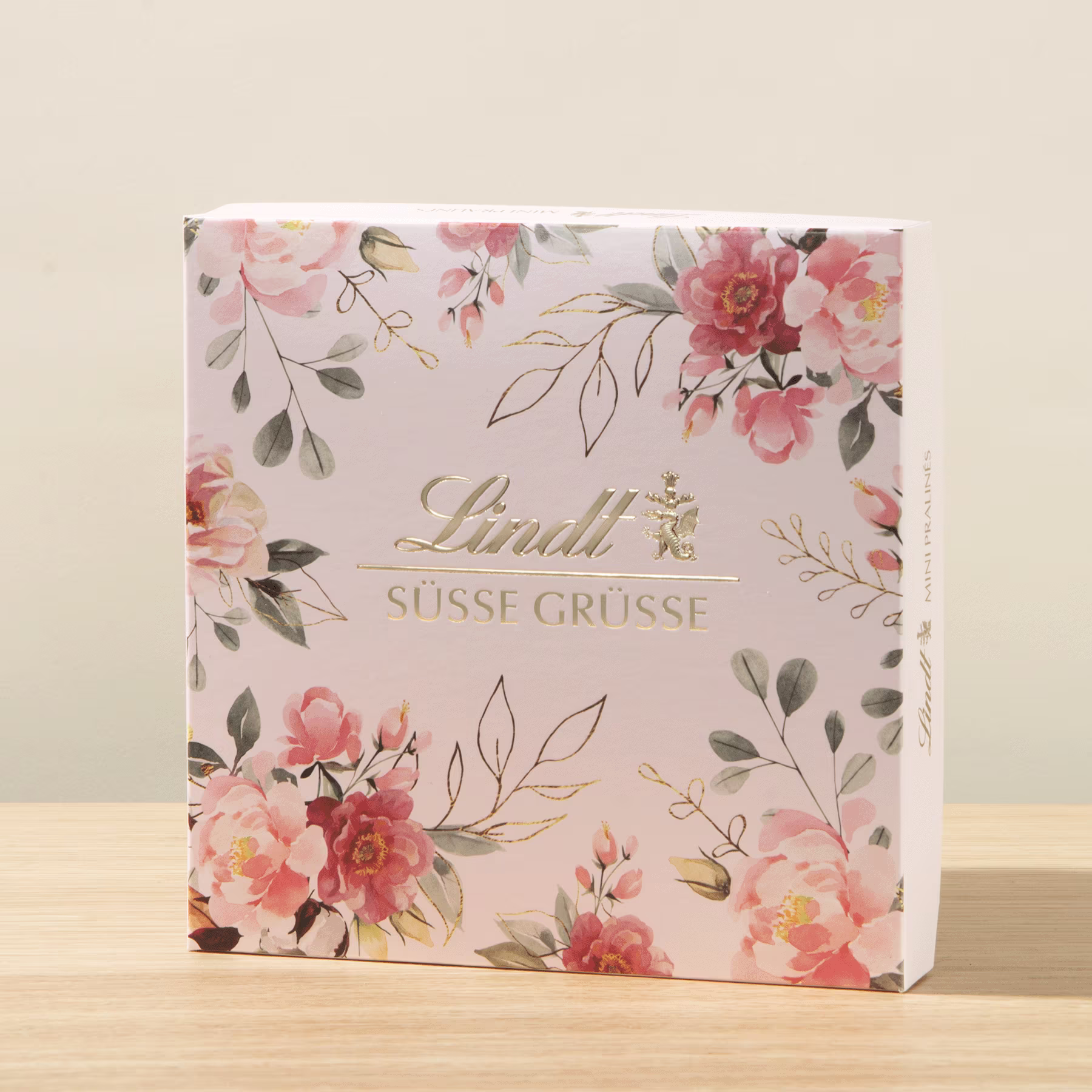 Eine quadratische Geschenkbox mit einem Blumenmuster aus rosa und weißen Blumen, Blättern und einem goldfarbenen Logo auf hellem Hintergrund. (Beschreibungstext KI-generiert)
