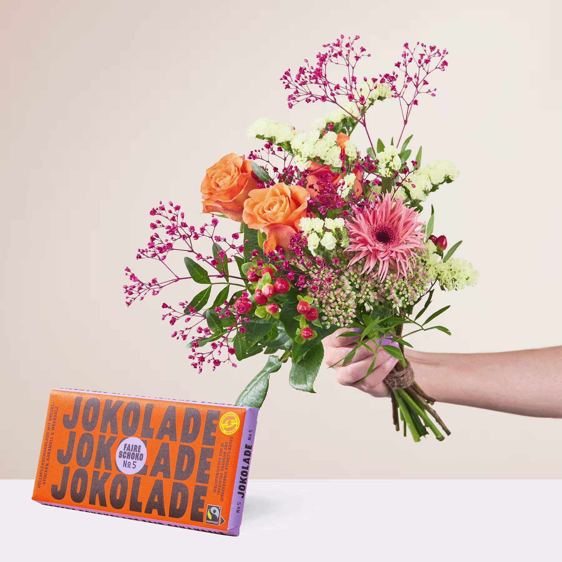 Eine Person hält einen leuchtend bunten Blumenstrauß, darunter orangefarbene Rosen, rosa Gerbera und Grünpflanzen, in der Hand. Im Vordergrund steht auf einer hellen Oberfläche eine Schachtel Jokolade-Schokoriegel. (Beschreibungstext KI-generiert)