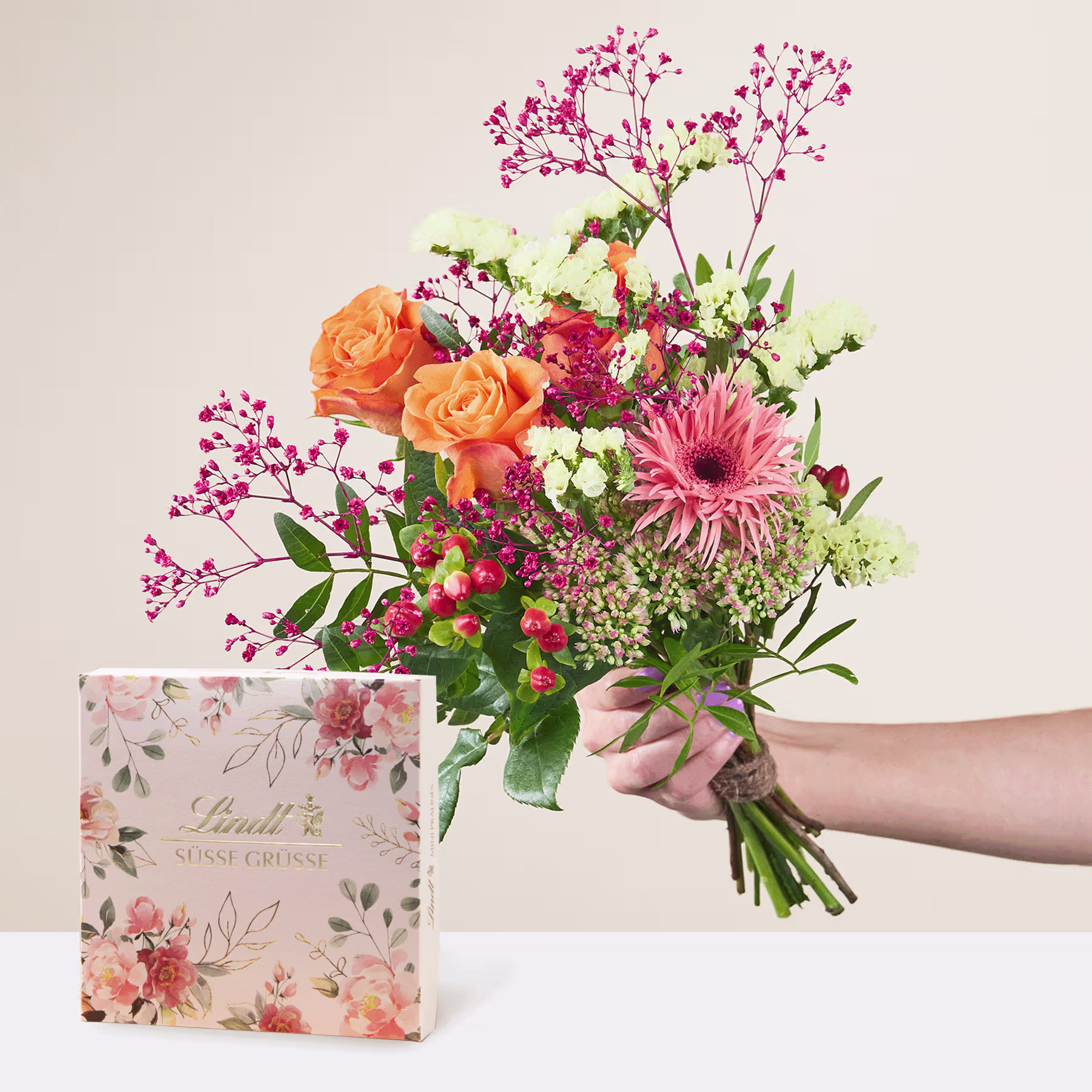 Ein lebendiger Strauß aus orangefarbenen Rosen, rosa Gerbera und anderen bunten Blumen, arrangiert in einem Strauß, begleitet von einer Geschenkbox mit Blumenmuster im Vordergrund. (Beschreibungstext KI-generiert)
