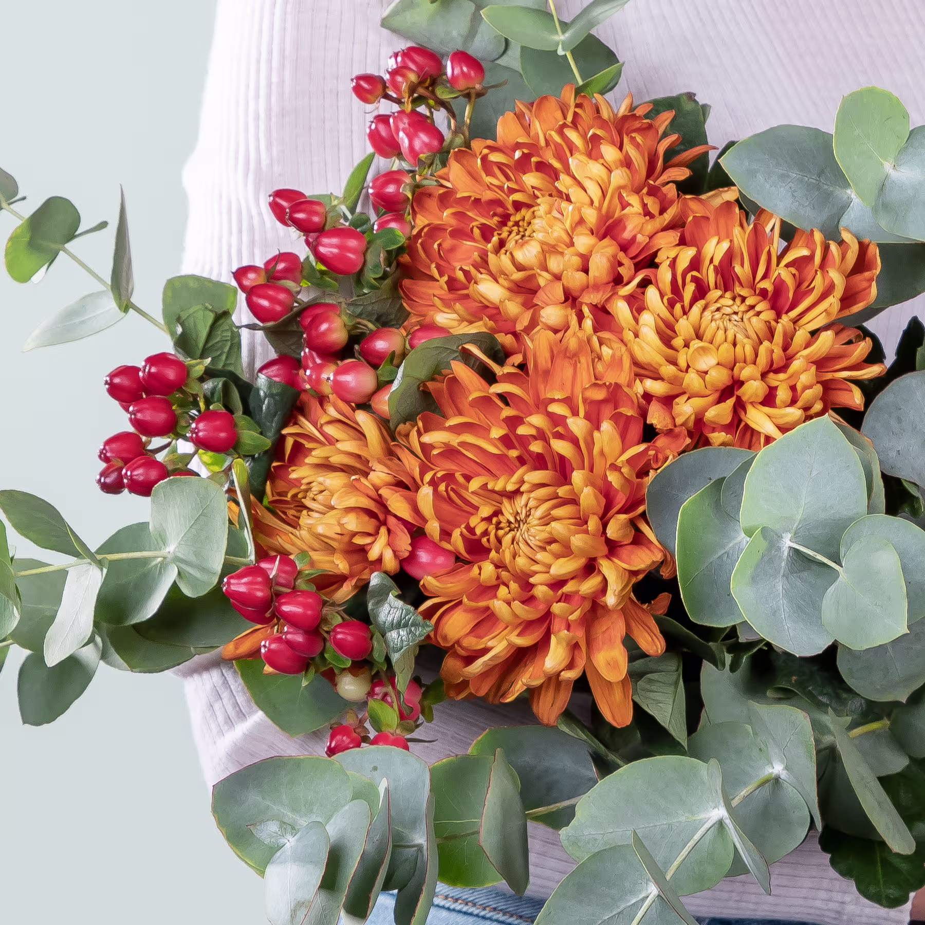 Ein lebendiger Strauß orangefarbener Chrysanthemen, umgeben von üppigem grünem Laub und roten Beeren, vor einem schlichten weißen Hintergrund. (Beschreibungstext KI-generiert)