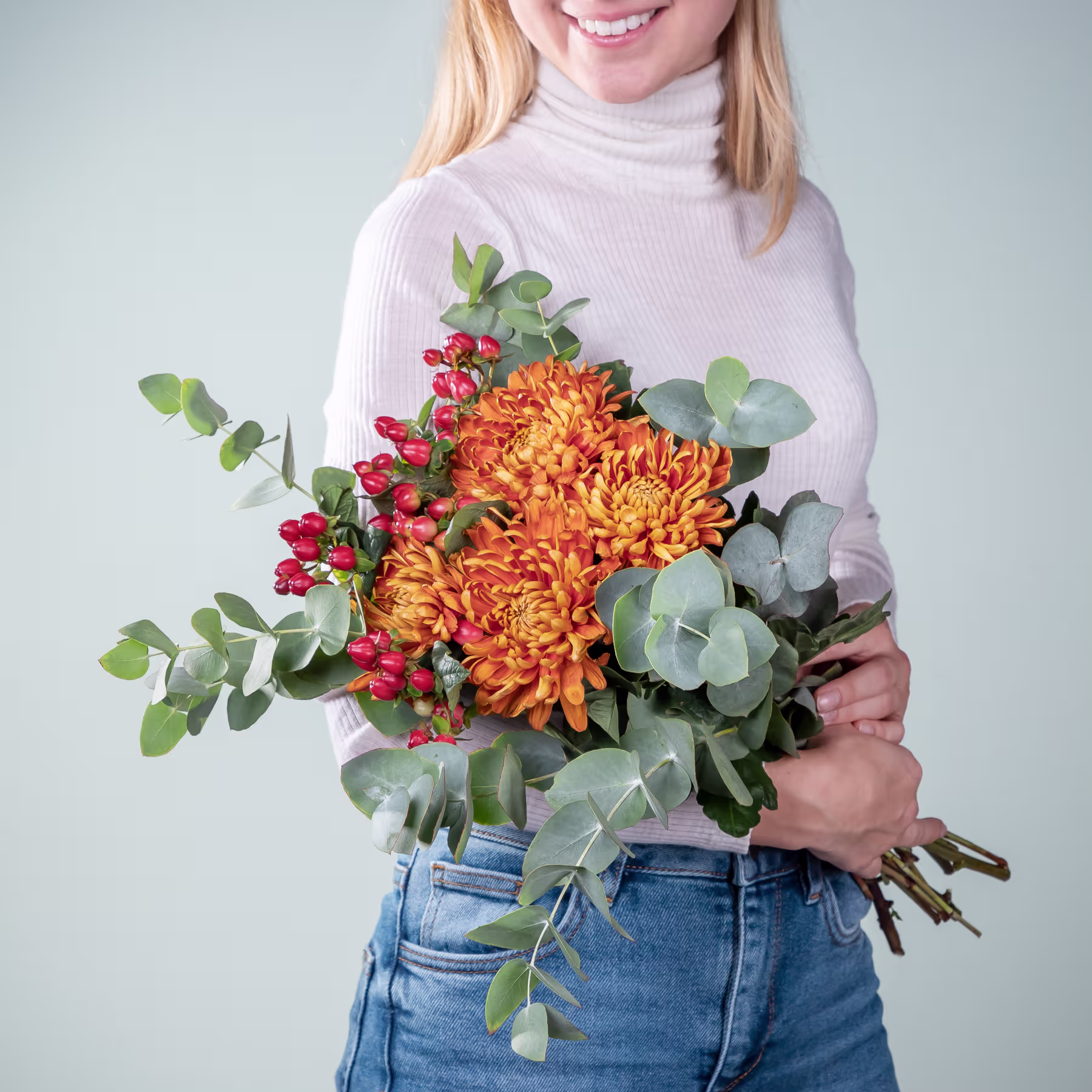Eine lächelnde Frau in einem weißen Pullover hält vor einem schlichten Hintergrund einen farbenfrohen Strauß orangefarbener Chrysanthemen und Grünpflanzen. (Beschreibungstext KI-generiert)