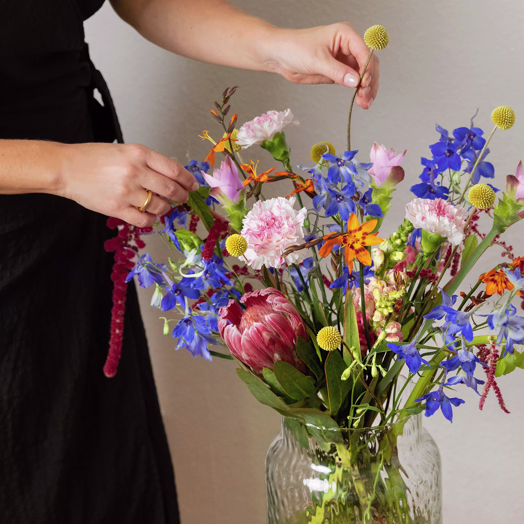 Ein lebendiger Strauß bunter Blumen, darunter Pfingstrosen, Gänseblümchen und Schwertlilien, steht in einer Glasvase vor einem schlichten Hintergrund. (Beschreibungstext KI-generiert)