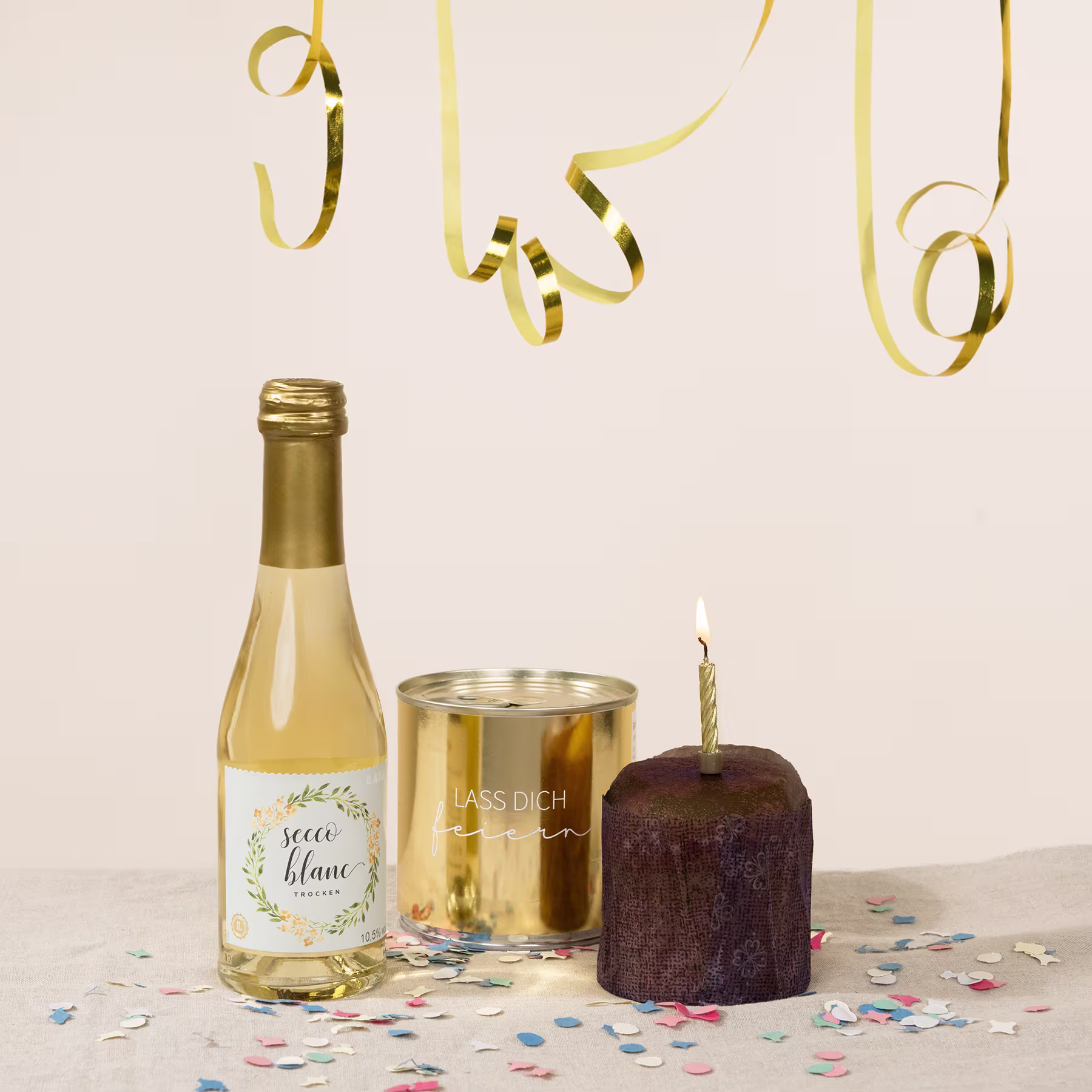 Eine Flasche Weißwein, eine goldene Blechdose und eine brennende Geburtstagskerze auf einem Tisch, umgeben von buntem Konfetti und sich kräuselnden Bändern. (Beschreibungstext KI-generiert)