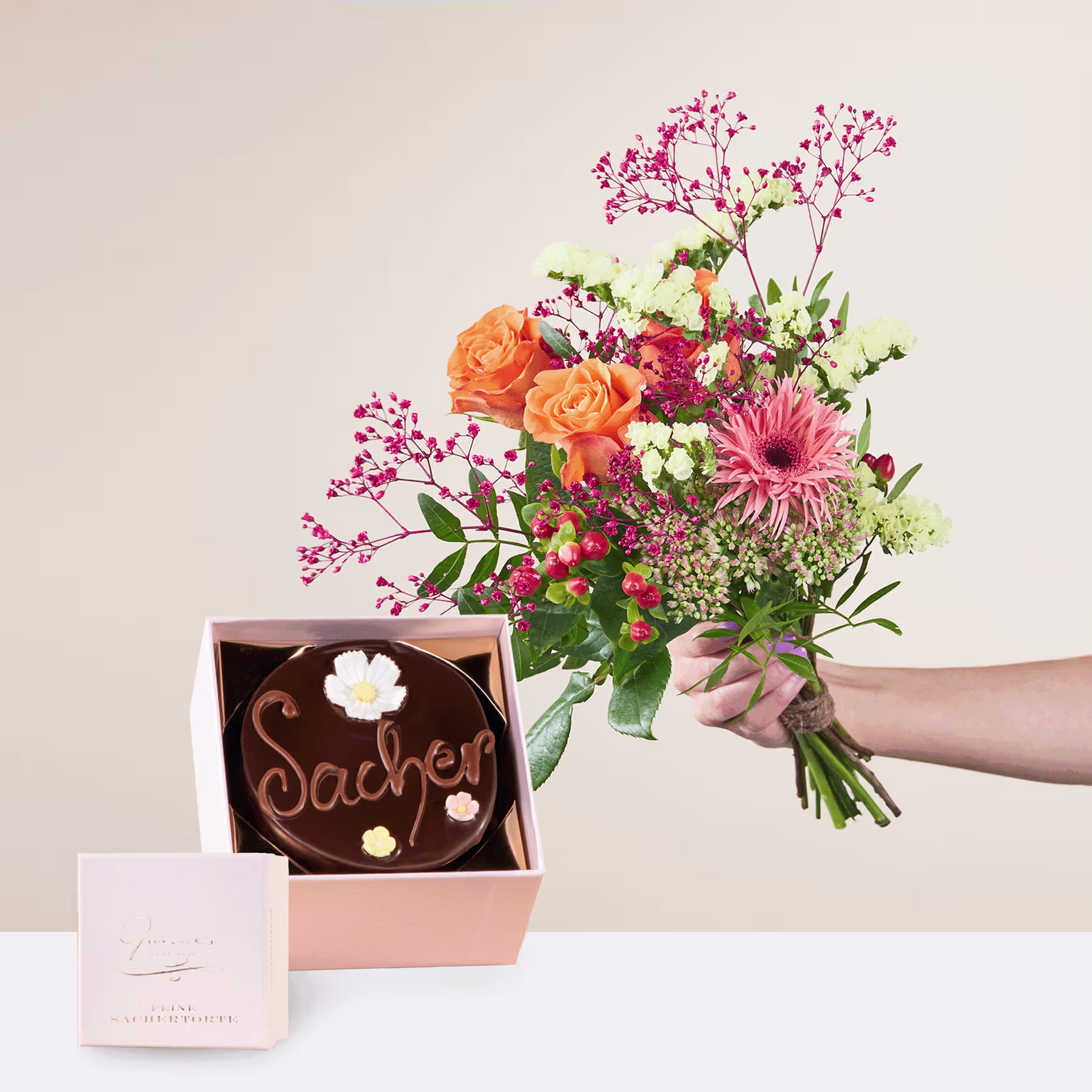 Eine Person hält einen leuchtend bunten Blumenstrauß, darunter orangefarbene Rosen und rosa Gerbera, in den Händen, zusammen mit einer Holzschachtel, die eine Schokoladenleckerei mit der Aufschrift „Sachet“ enthält. (Beschreibungstext KI-generiert)