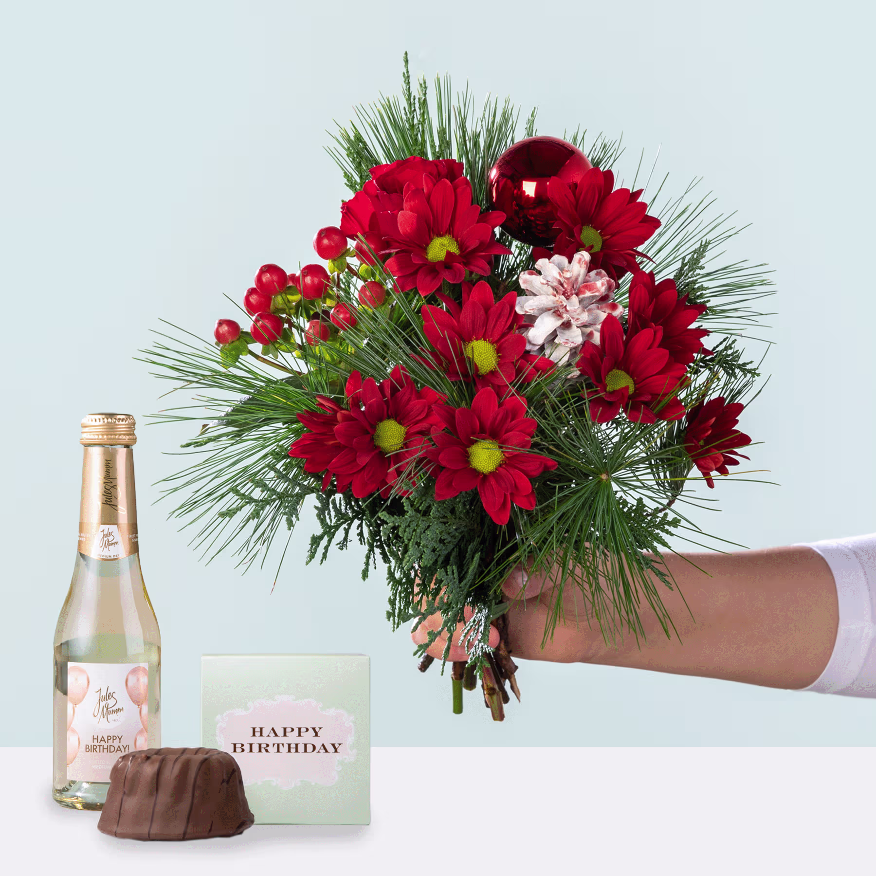 Ein farbenfroher Strauß roter und rosa Blumen, darunter Chrysanthemen und Tannenzweige, wird vor einem schlichten Hintergrund in einer Hand gehalten, begleitet von einer Flasche Wein und einer Geburtstagskarte. (Beschreibungstext KI-generiert)