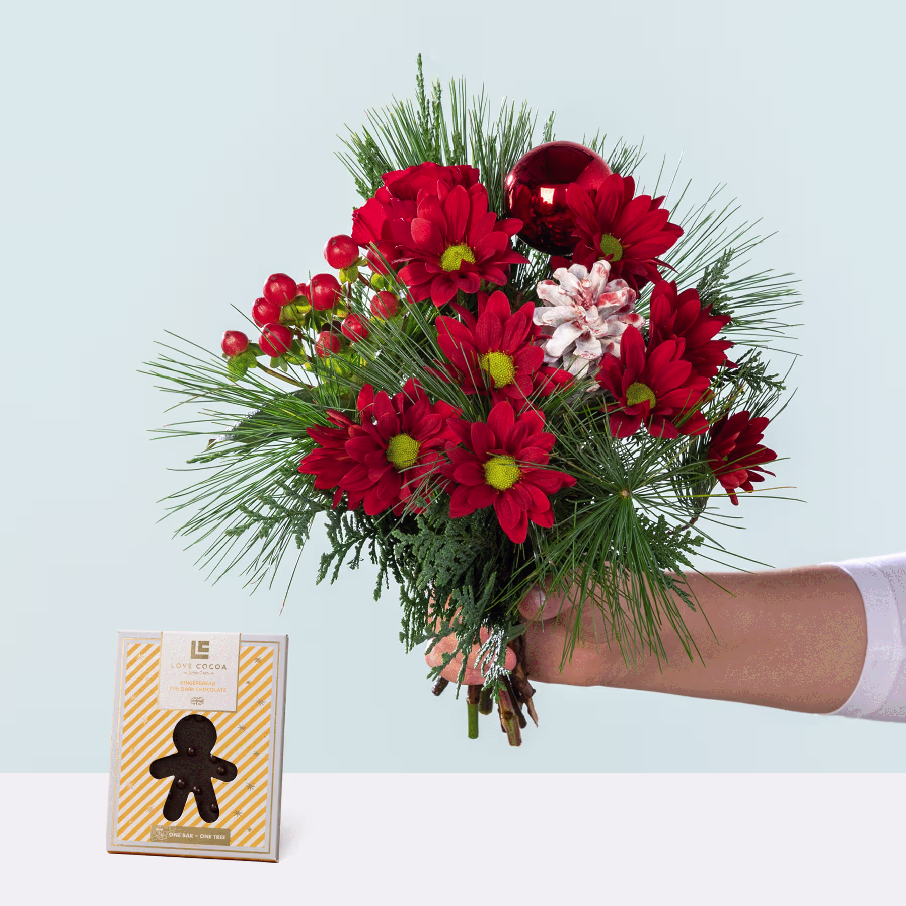 Ein farbenfroher Strauß roter und rosa Blumen, darunter Chrysanthemen und Tannenzweige, wird vor einem schlichten Hintergrund in einer Hand gehalten, im Vordergrund steht eine kleine Geschenkbox in Form eines Lebkuchenmanns. (Beschreibungstext KI-generiert)