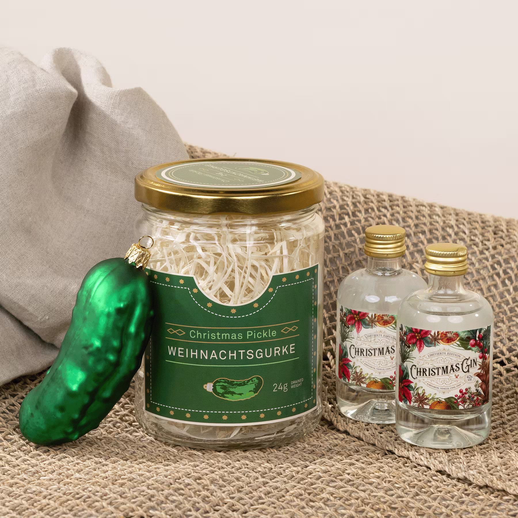 Auf einer Juteoberfläche stehen ein Glas mit eingelegtem Gemüse, eine grüne Essiggurke und zwei kleine Glasflaschen mit festlichen Weihnachtsetiketten. (Beschreibungstext KI-generiert)