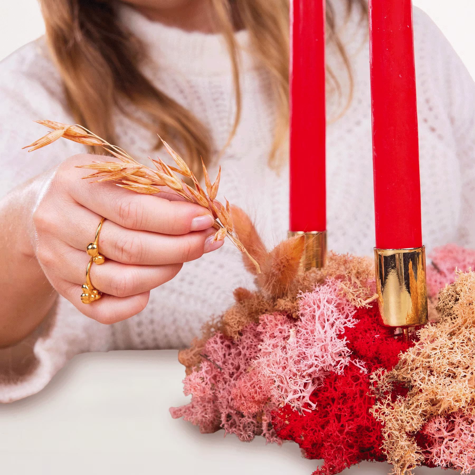 Die Hände einer Person halten ein Bündel Weizenhalme und eine rote Kerze vor einem Hintergrund aus rosa und roten Blumenarrangements. (Beschreibungstext KI-generiert)