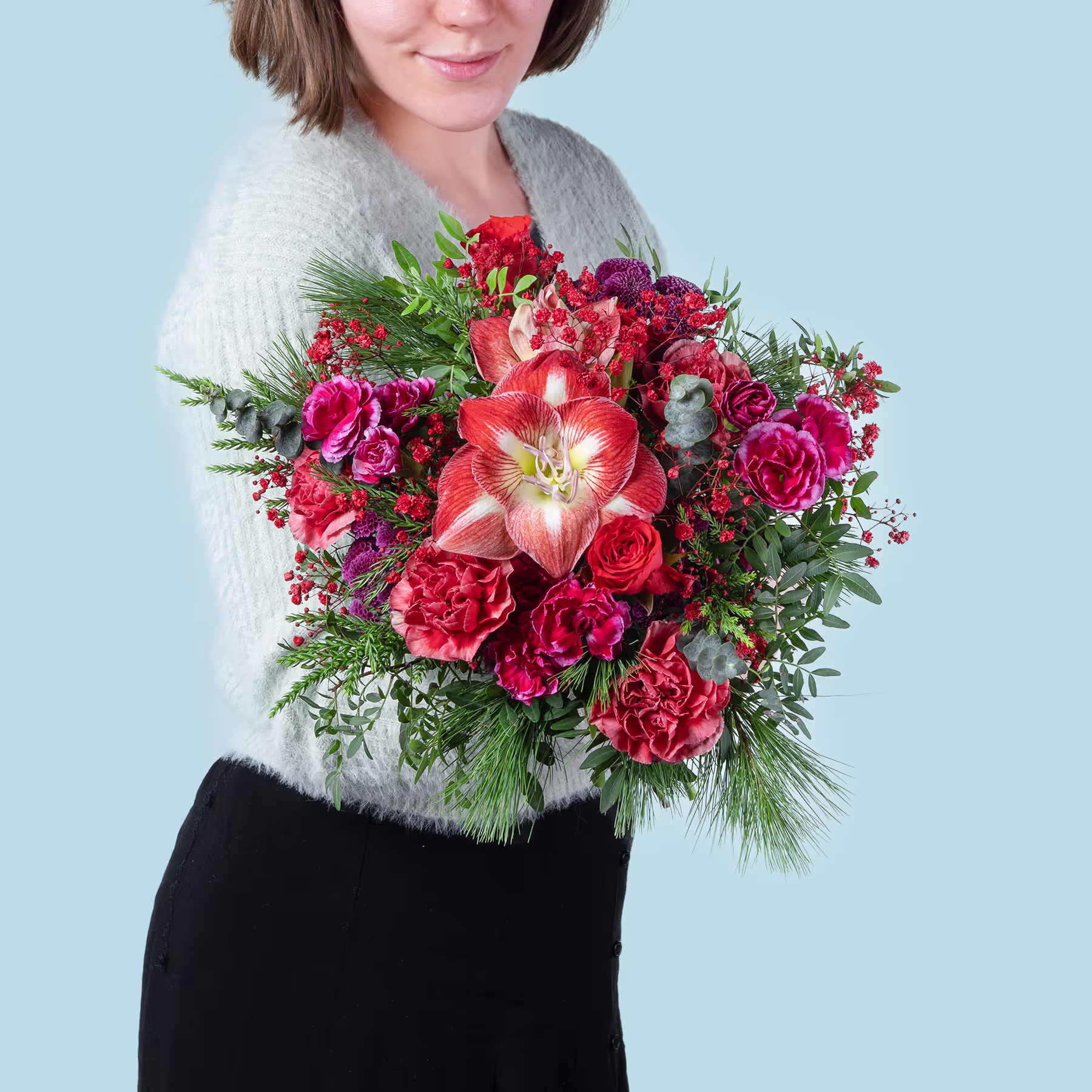 Eine Frau in einem grauen Pullover hält vor einem hellblauen Hintergrund einen farbenfrohen Strauß roter und rosa Blumen, darunter Rosen und Lilien. (Beschreibungstext KI-generiert)