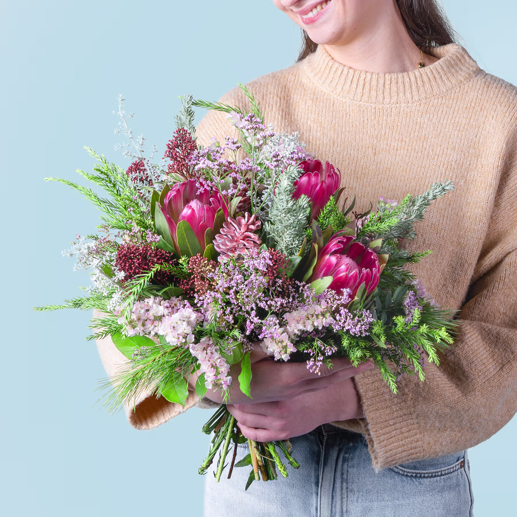 Eine Frau in einem beigen Pullover hält einen farbenfrohen Strauß aus rosa Protea-Blüten, violetten und weißen Wildblumen und üppigem Grün vor einem schlichten blauen Hintergrund. (Beschreibungstext KI-generiert)