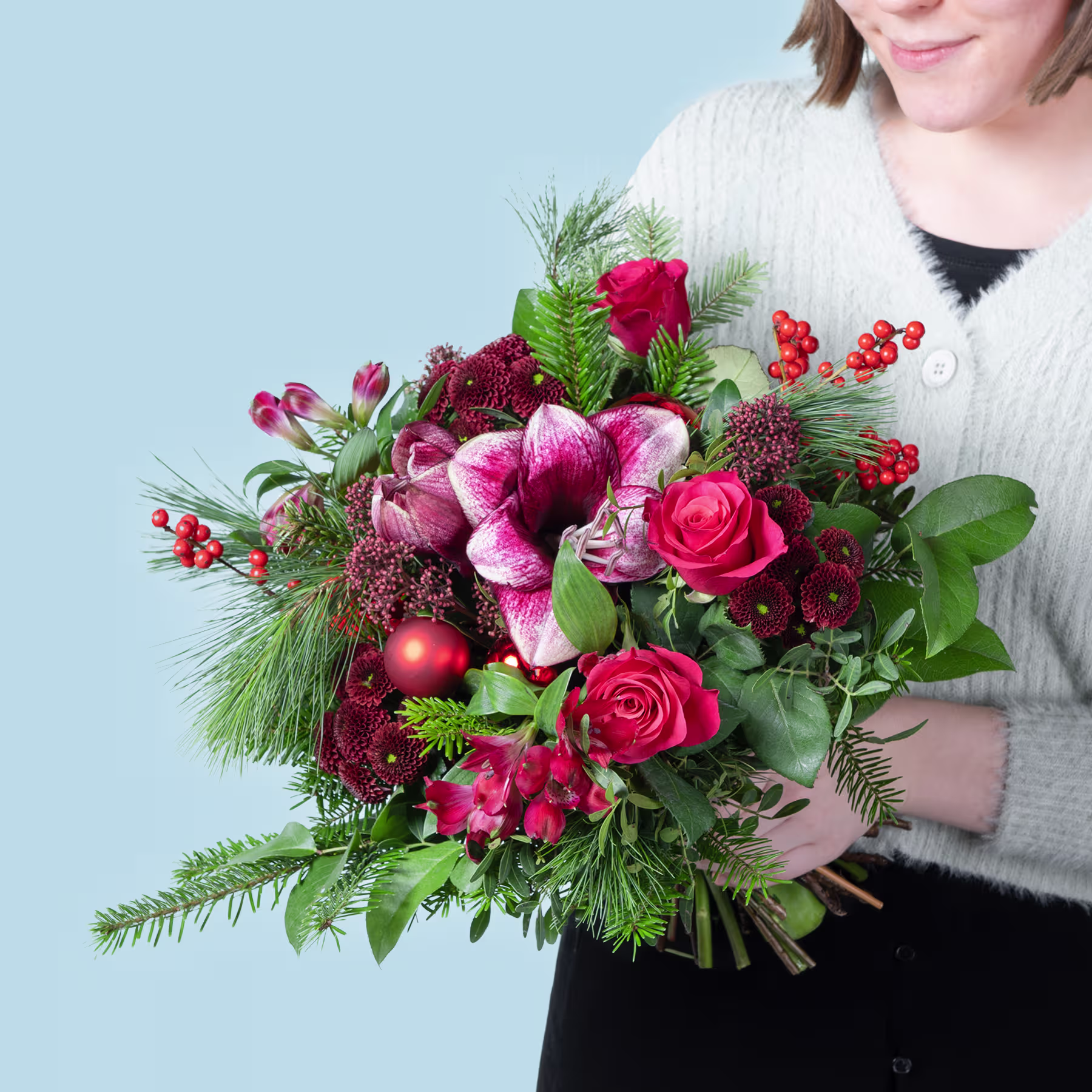 Ein leuchtender Strauß roter und rosa Blumen, darunter Rosen, Orchideen und Beeren, gehalten von einer Person in einem weißen Pullover vor einem schlichten blauen Hintergrund. (Beschreibungstext KI-generiert)