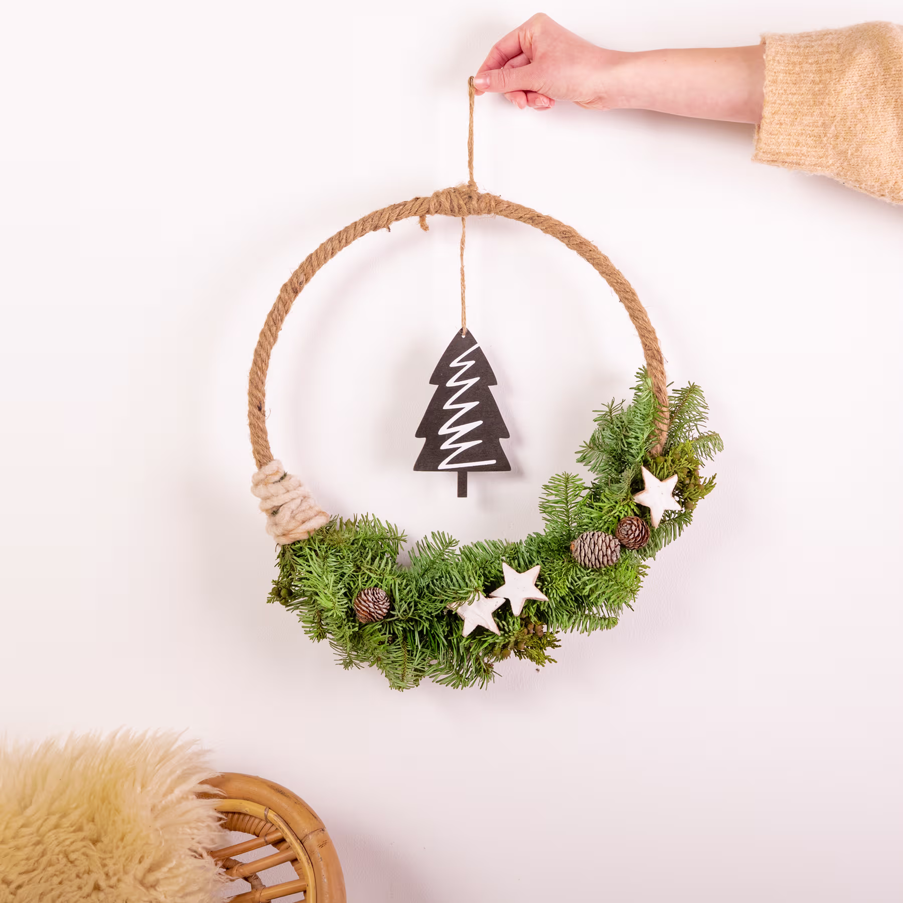 Eine Hand hängt einen dekorativen Weihnachtskranz aus immergrünen Zweigen, Sternen und einem hölzernen Baumschmuck an eine weiße Wand. (Beschreibungstext KI-generiert)