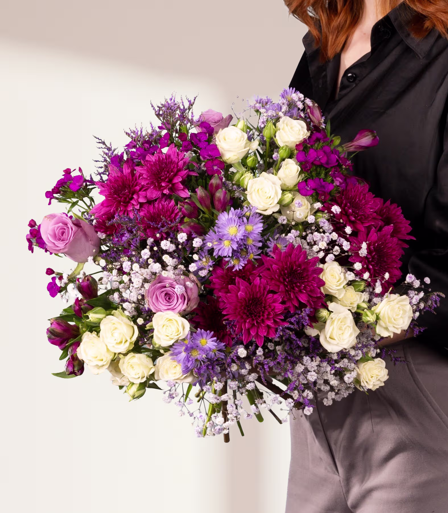 Ein farbenfroher Strauß verschiedener Blumen, darunter rosa Rosen, lila Gänseblümchen und weiße Nelken, gehalten von einer Person in einer schwarzen Jacke vor einem schlichten Hintergrund. (Beschreibungstext KI-generiert)