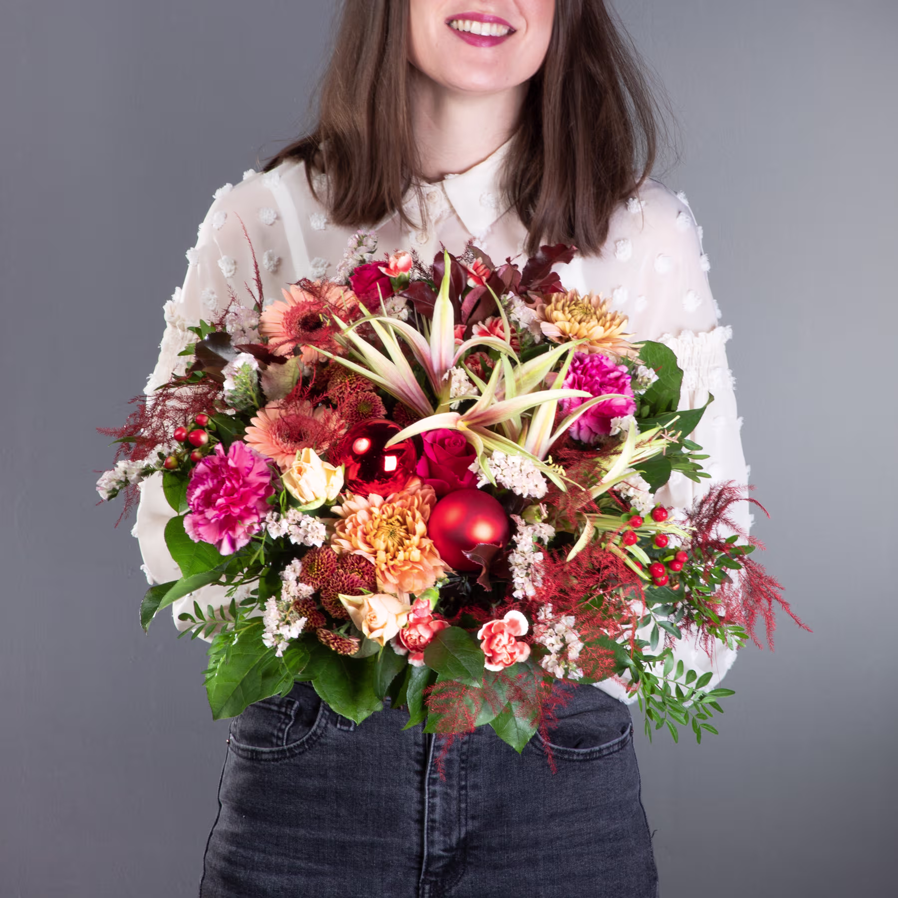 Eine lächelnde junge Frau hält vor einem schlichten grauen Hintergrund einen farbenfrohen Strauß herbstlicher Blumen, darunter rote, rosa und gelbe Blüten. (Beschreibungstext KI-generiert)