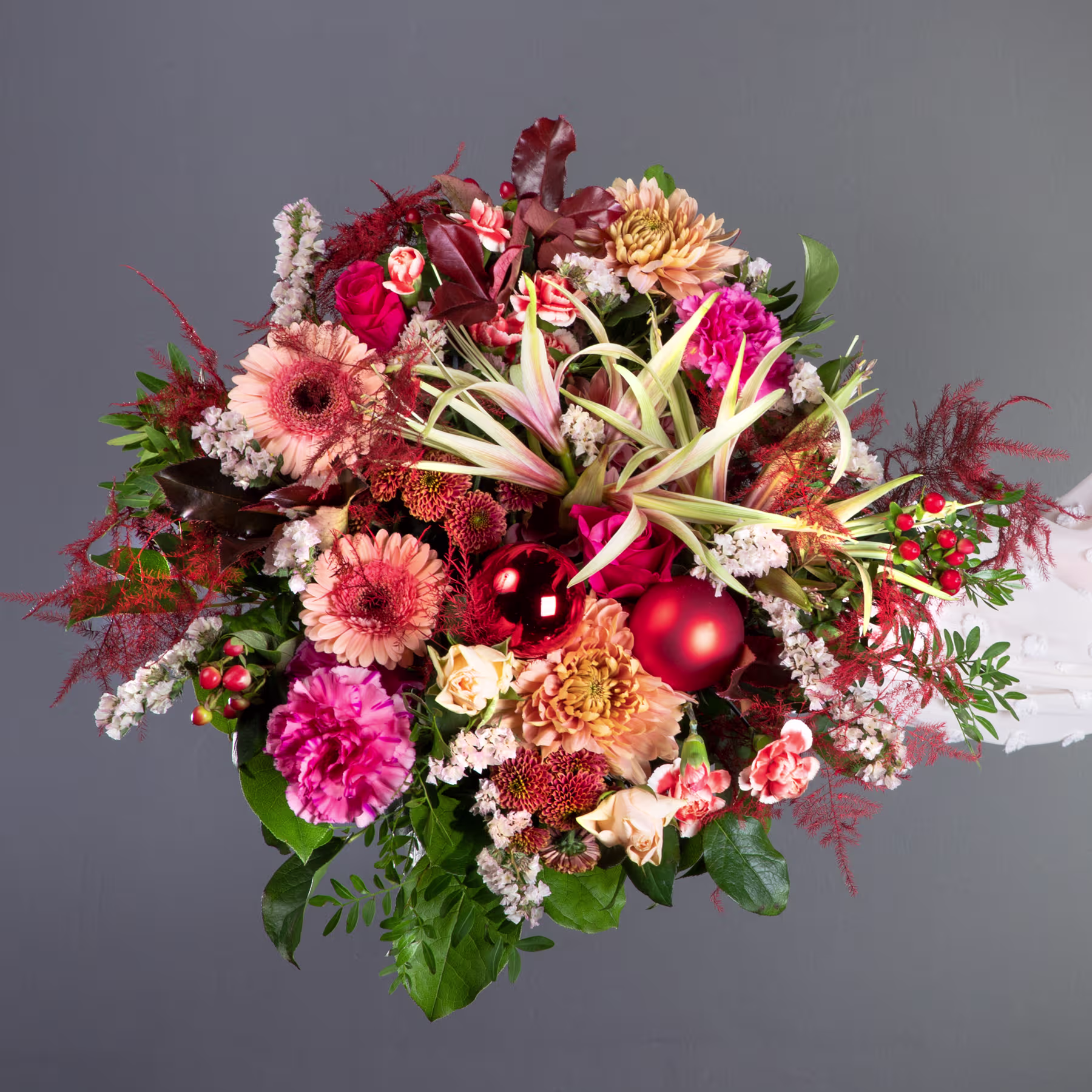 Ein lebendiger und üppiger Strauß verschiedener bunter Blumen, darunter rote, rosa und gelbe Blüten sowie Grünpflanzen, vor einem schlichten grauen Hintergrund. (Beschreibungstext KI-generiert)