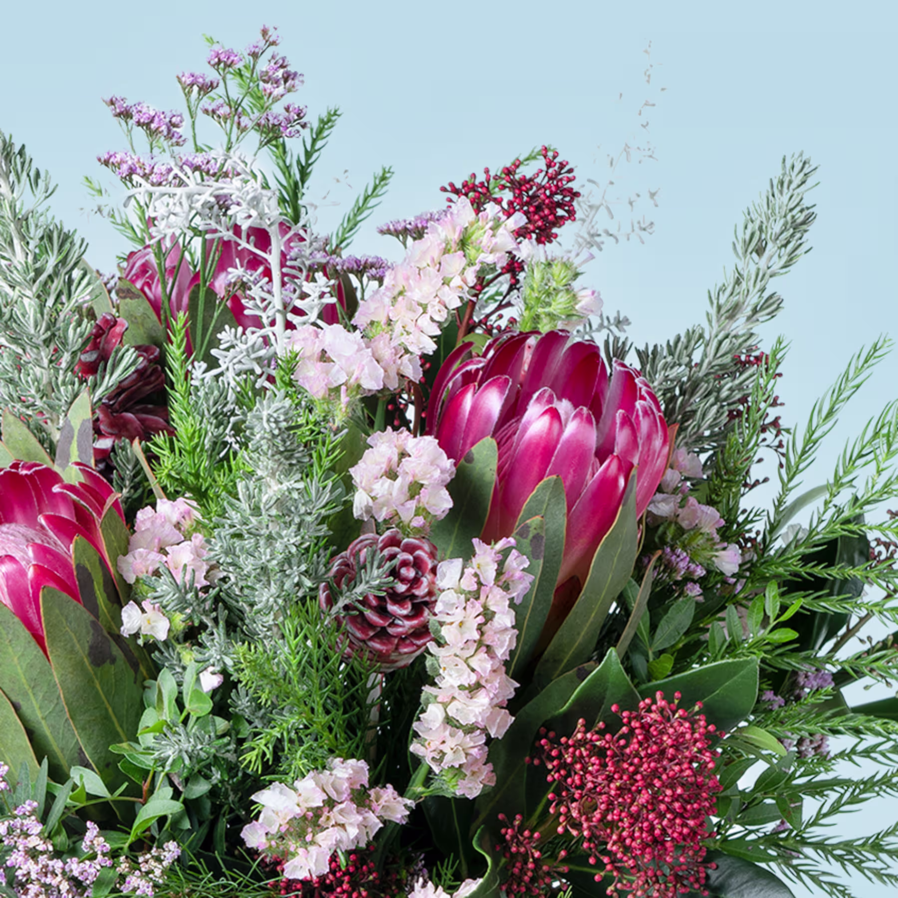 Ein lebendiger Strauß verschiedener Blumen, darunter große rosa Proteablüten, zarte weiße Blumen und üppiges Grün vor einem sanften blauen Hintergrund. (Beschreibungstext KI-generiert)
