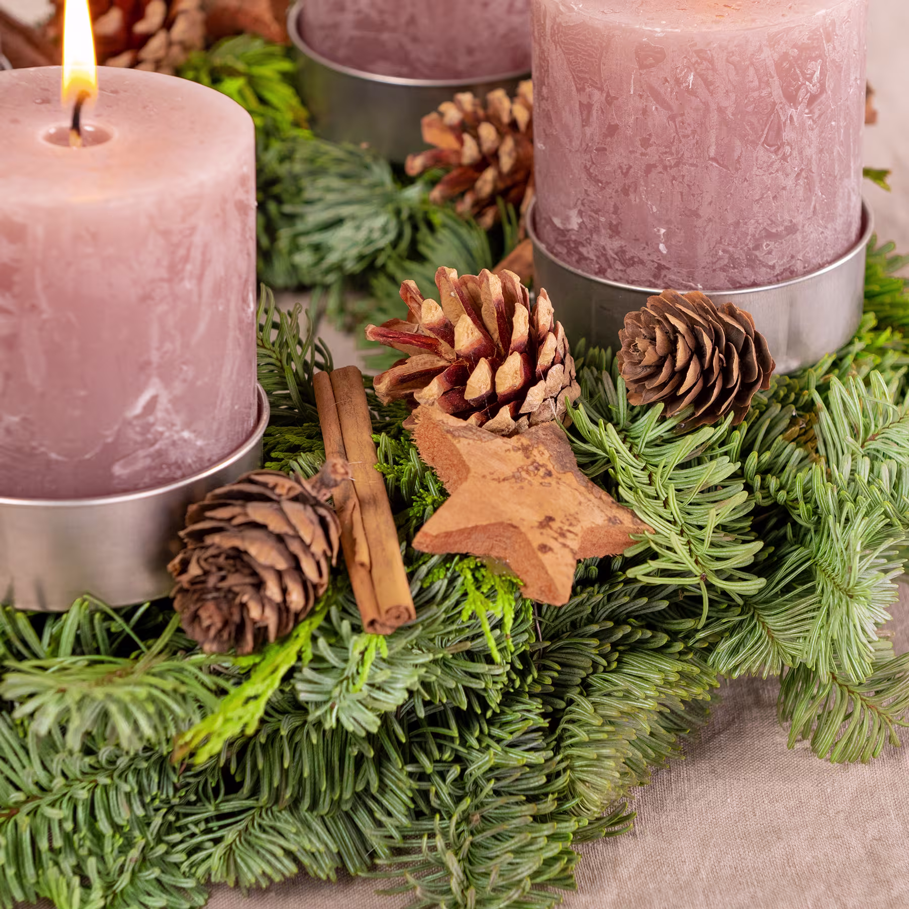 Das Bild zeigt ein festliches Weihnachtsarrangement mit brennenden Kerzen, Tannenzapfen und einer hölzernen Sterndekoration, eingebettet zwischen üppigen immergrünen Zweigen. (Beschreibungstext KI-generiert)