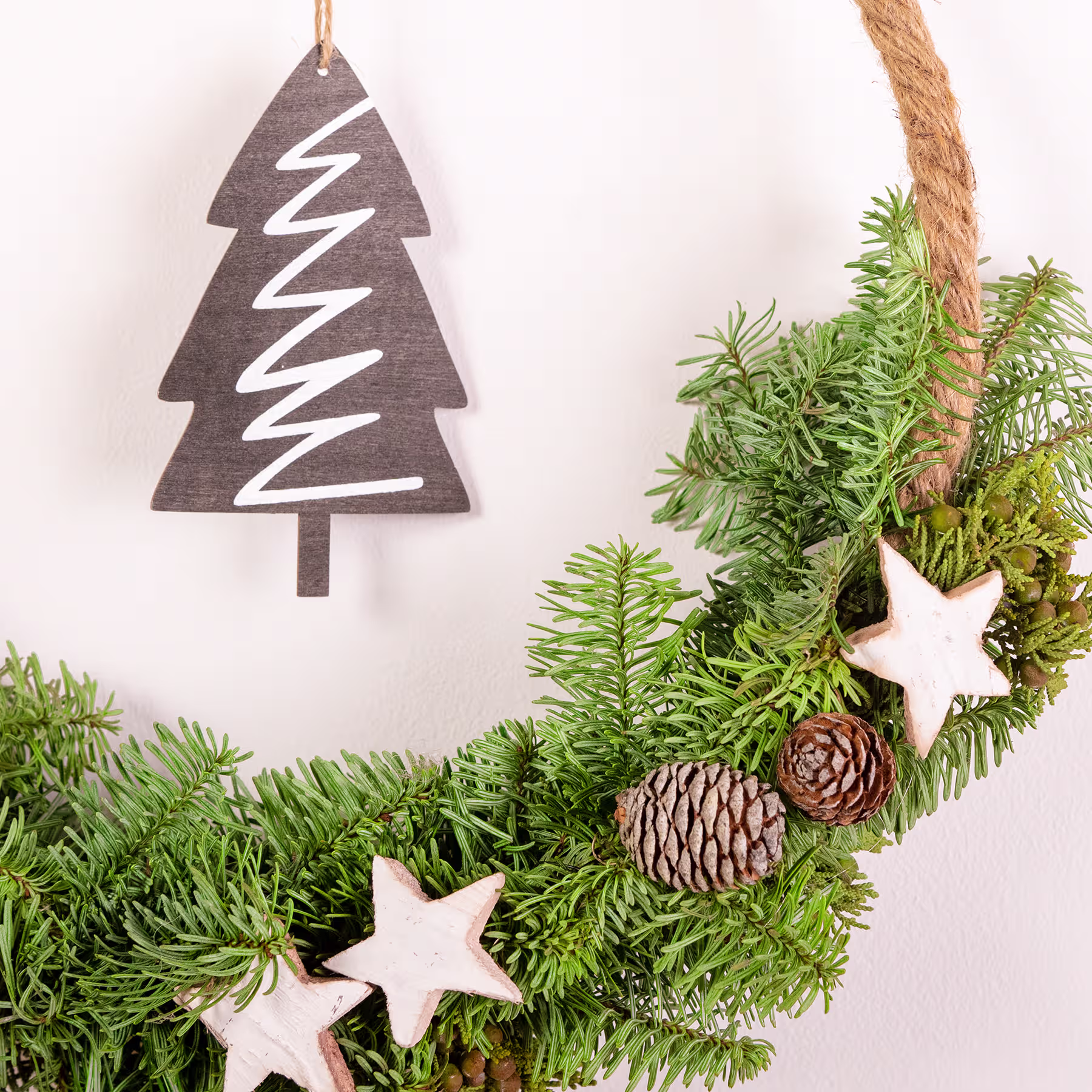 Ein festlicher Weihnachtskranz aus Tannenzweigen, Tannenzapfen und hölzernen Sterndekorationen, im Hintergrund hängt ein baumförmiges Holzornament. (Beschreibungstext KI-generiert)