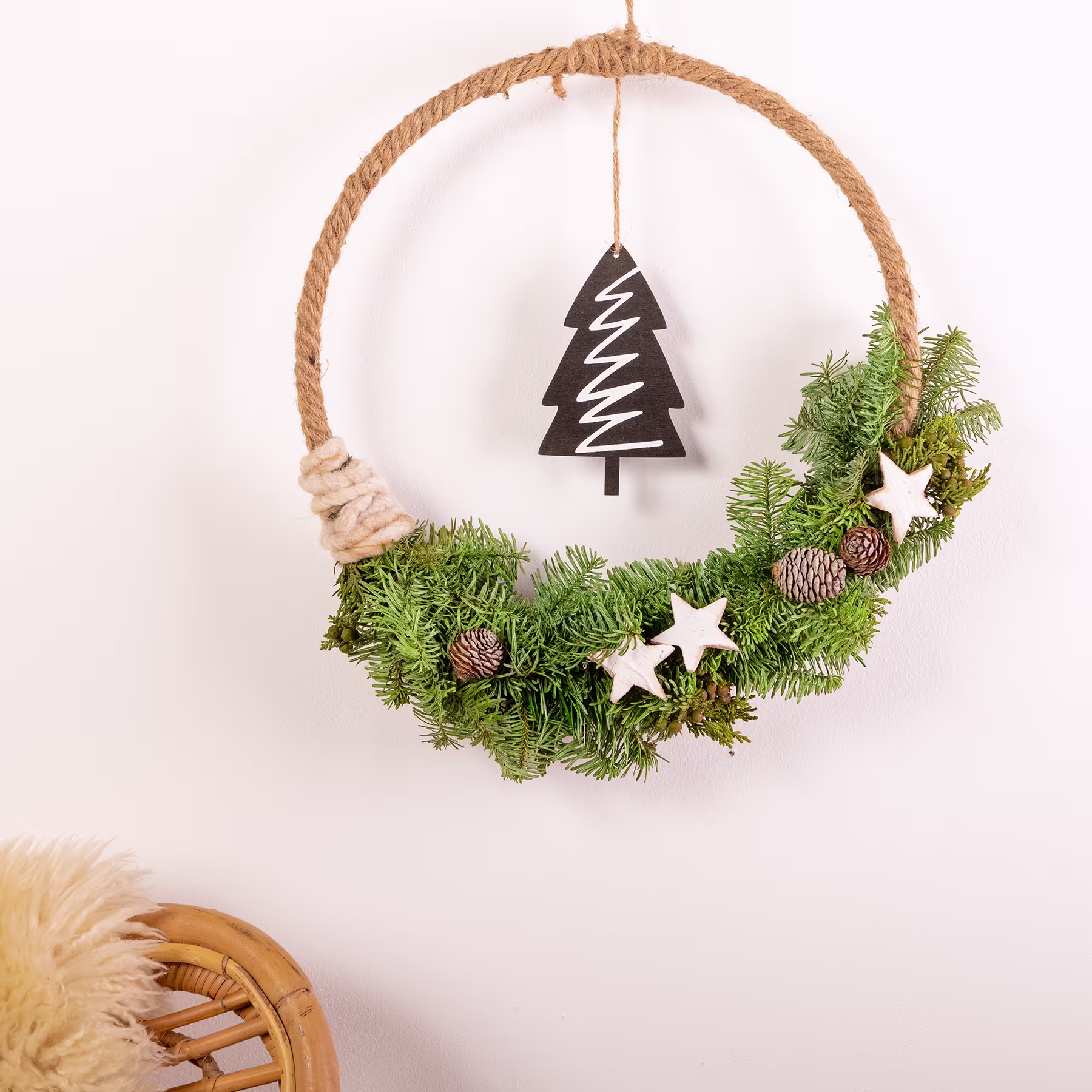 Ein rustikaler Weihnachtskranz aus Tannenzweigen, Tannenzapfen und einem hölzernen Baumschmuck, der vor einem schlichten weißen Hintergrund hängt. (Beschreibungstext KI-generiert)