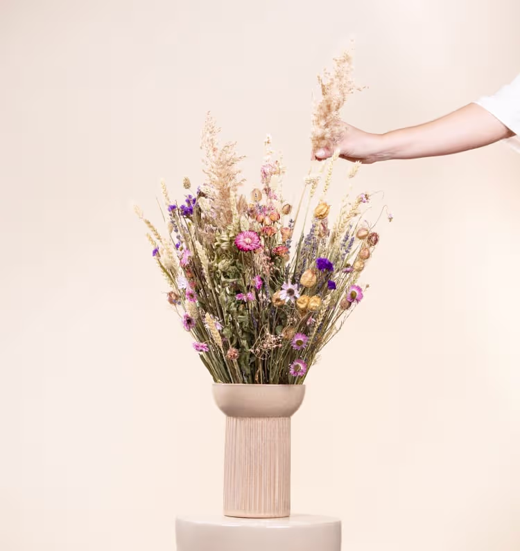 Eine Hand arrangiert einen Strauß bunter Wildblumen in einer Holzvase vor einem schlichten, hellen Hintergrund. (Beschreibungstext KI-generiert)