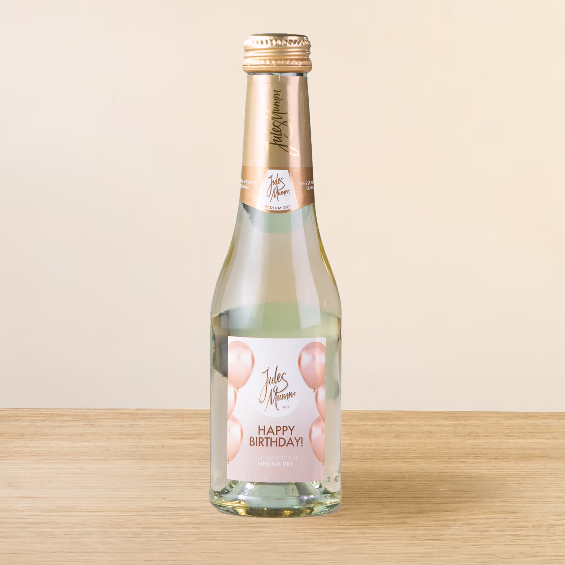 Eine Glasflasche Sekt oder Champagner mit dem Etikett „Happy Birthday!“ auf einer Holzoberfläche vor hellem Hintergrund. (Beschreibungstext KI-generiert)