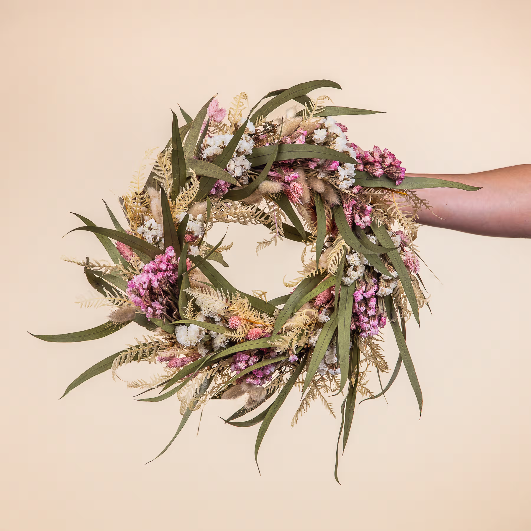 Ein handgefertigter Blumenkranz aus verschiedenen rosa und weißen Blumen, Blättern und Grünpflanzen vor einem neutralen beigen Hintergrund. (Beschreibungstext KI-generiert)