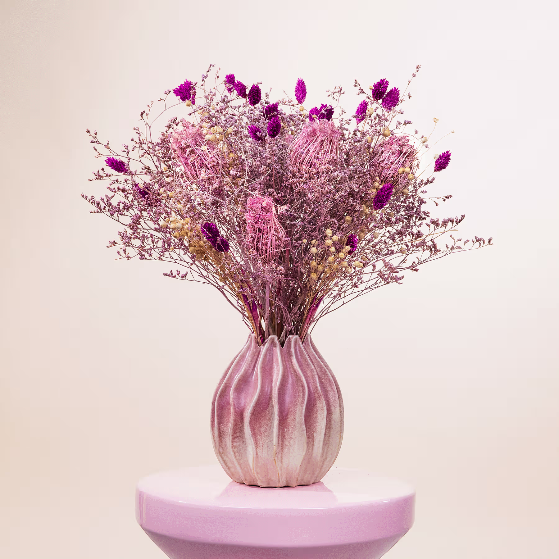 Ein farbenfroher Strauß getrockneter violetter und rosafarbener Blumen in einer zartrosa Vase steht auf einer blassrosa Fläche vor einem neutralen Hintergrund. (Beschreibungstext KI-generiert)