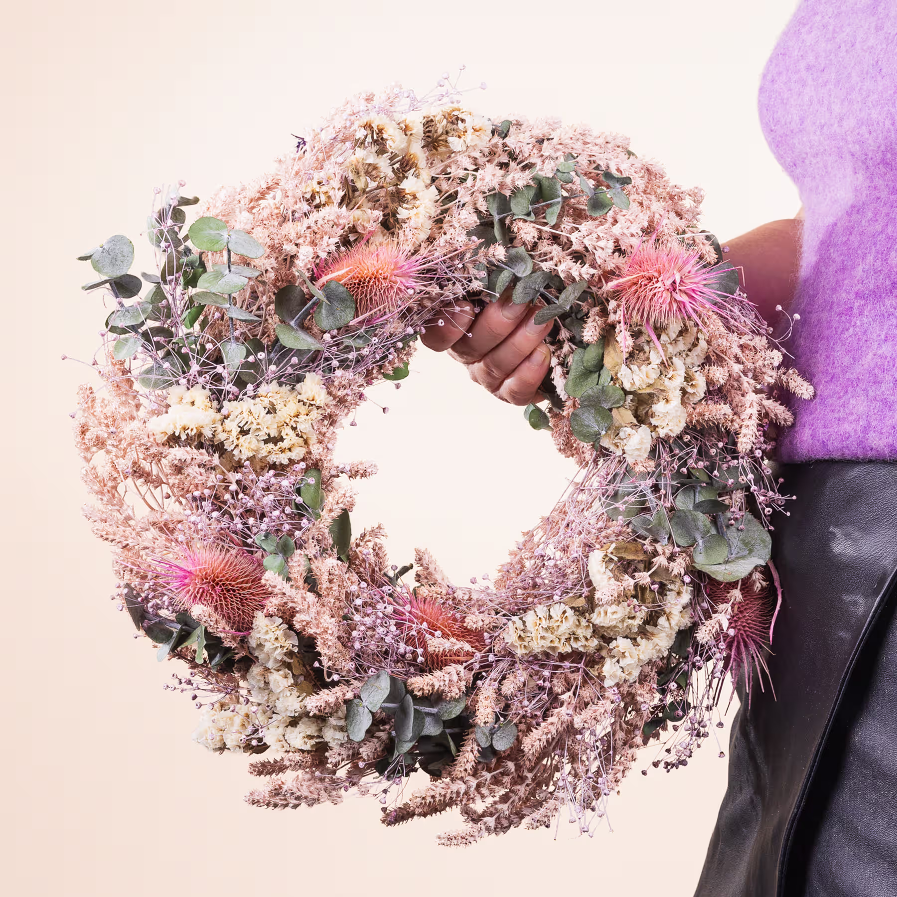 Ein lebendiger und komplizierter Blumenkranz, geschmückt mit einer Vielzahl zarter Blüten in Rosa-, Weiß- und Grüntönen, wird im Vordergrund vor einem neutralen Hintergrund gehalten. (Beschreibungstext KI-generiert)