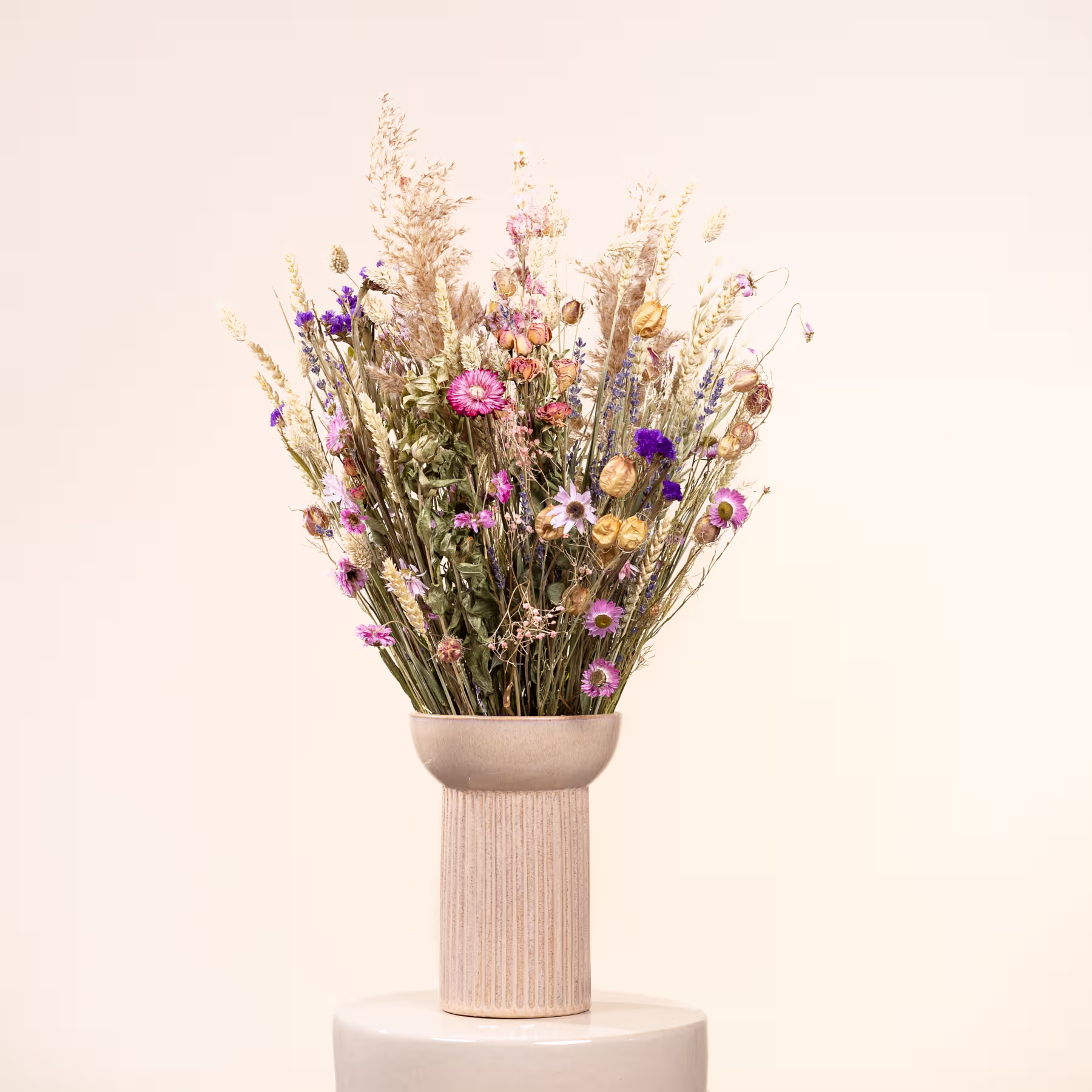 Ein Strauß leuchtender Trockenblumen, darunter rosa und violette Blüten, steht hoch in einer einfachen weißen Keramikvase vor einem schlichten, hellen Hintergrund. (Beschreibungstext KI-generiert)