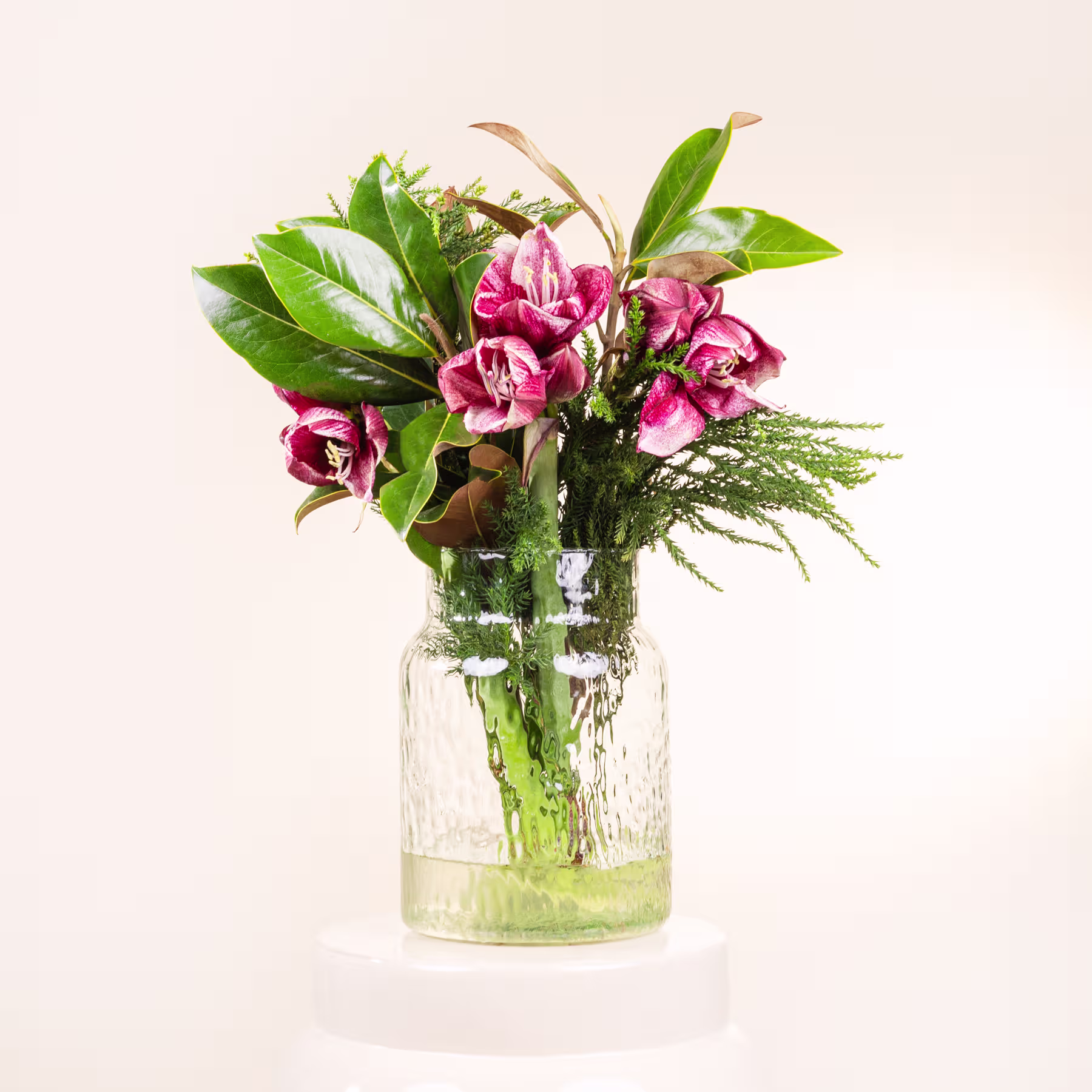 Ein Strauß leuchtend rosa Blumen, darunter Nelken und Grünpflanzen, wird in einer klaren Glasvase vor einem schlichten, hellen Hintergrund präsentiert. (Beschreibungstext KI-generiert)