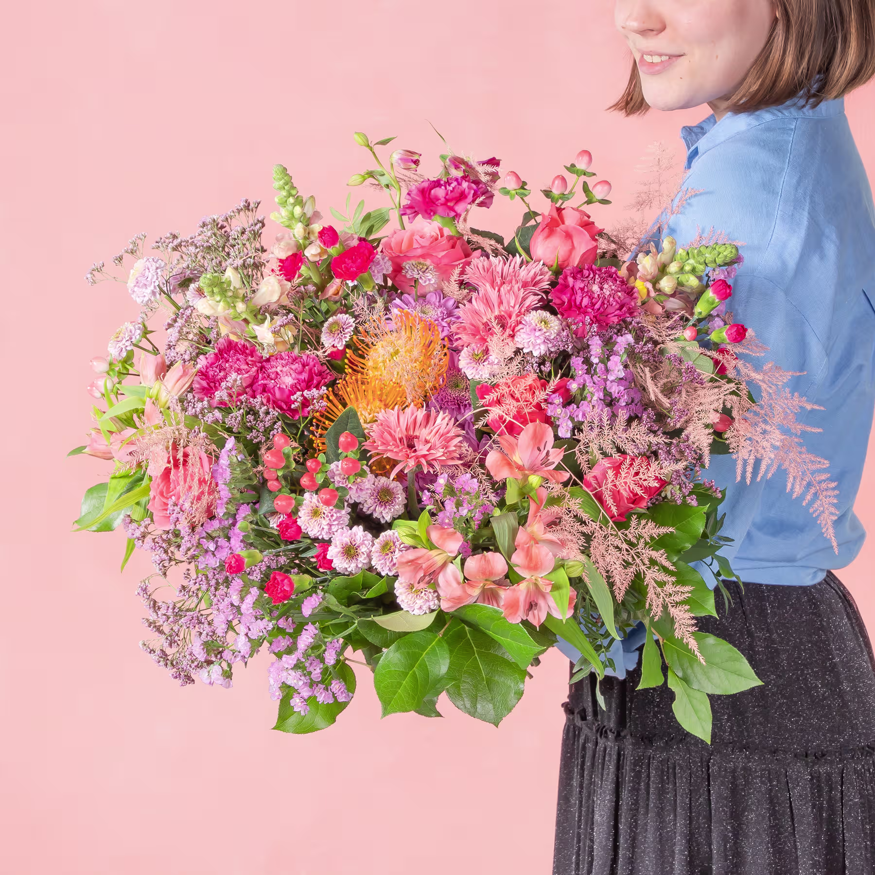 Ein leuchtender Strauß bunter Blumen, darunter rosa Nelken, gelbe Gänseblümchen und lila Flieder, wird von einer Person gehalten, die ein blaues Jeanshemd vor einem zartrosa Hintergrund trägt. (Beschreibungstext KI-generiert)