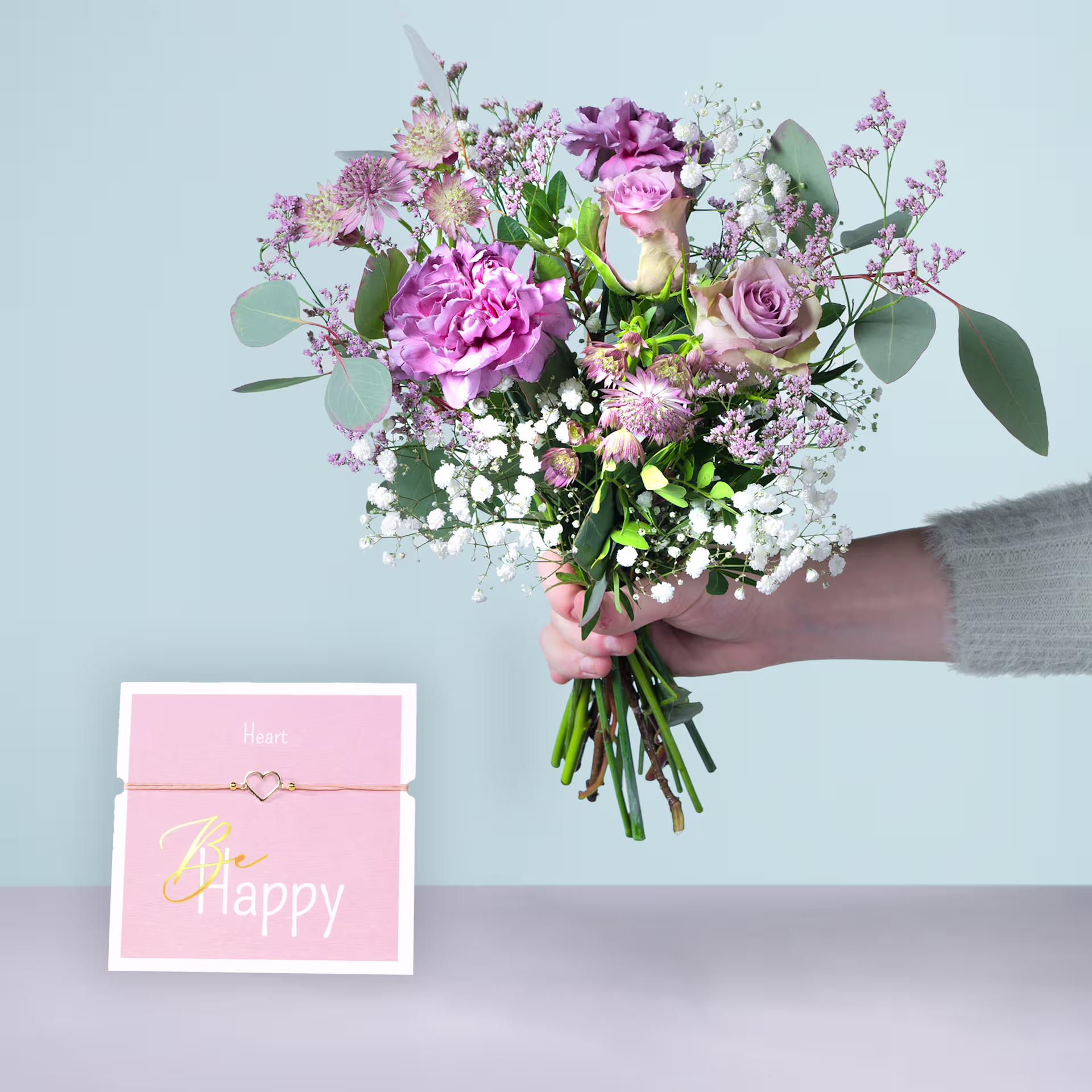 Eine Person hält einen farbenfrohen Strauß rosa und weißer Blumen, darunter Pfingstrosen und Rosen, in den Händen, begleitet von einer rosa Geschenkbox mit der Aufschrift „Happy“. (Beschreibungstext KI-generiert)