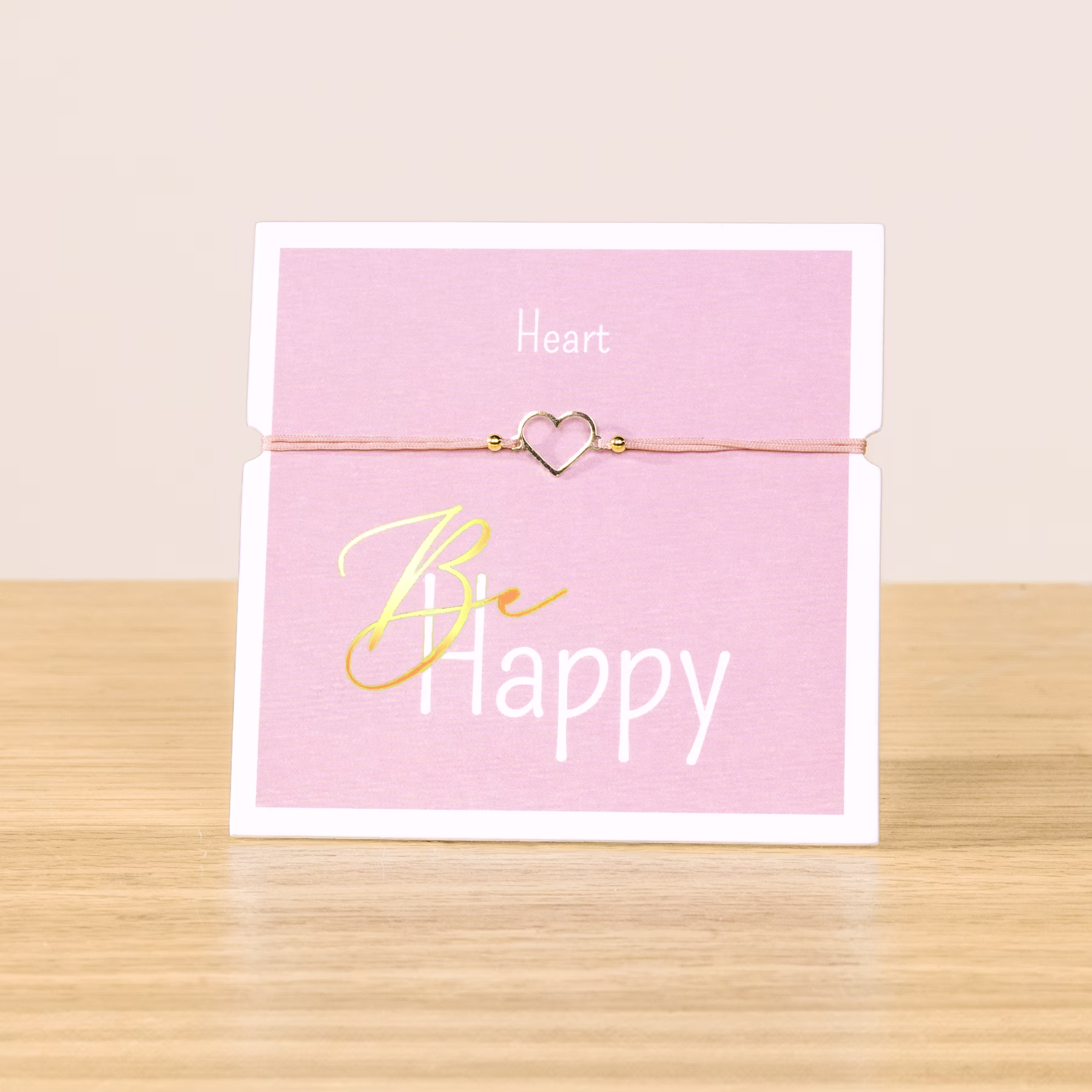Auf einer Holzoberfläche liegt eine rosa Karte mit dem Text „Be happy“ und einem herzförmigen Anhänger an einer Schnur. (Beschreibungstext KI-generiert)