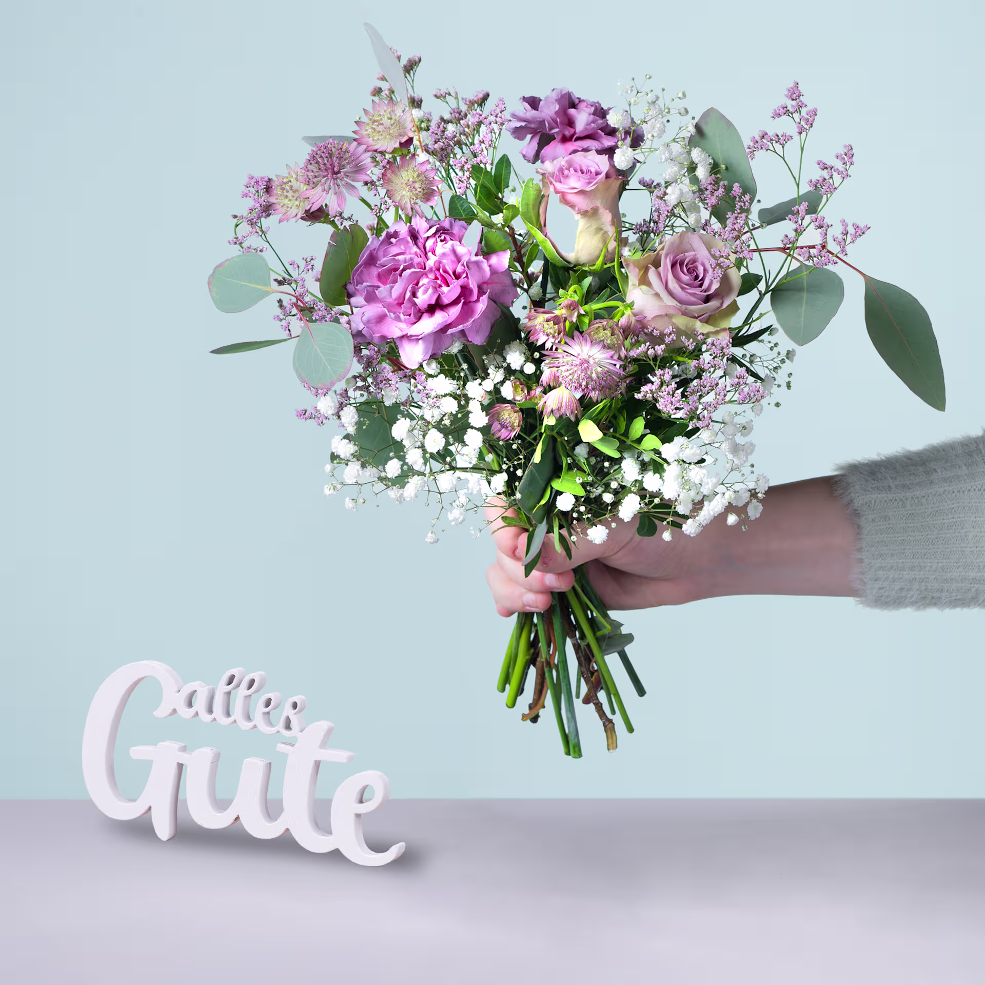 Ein farbenfroher Strauß rosa und weißer Blumen, darunter Pfingstrosen und Rosen, wird vor einem hellblauen Hintergrund in den Händen einer Person gehalten, im Vordergrund ist das Wort „Gute“ zu sehen. (Beschreibungstext KI-generiert)